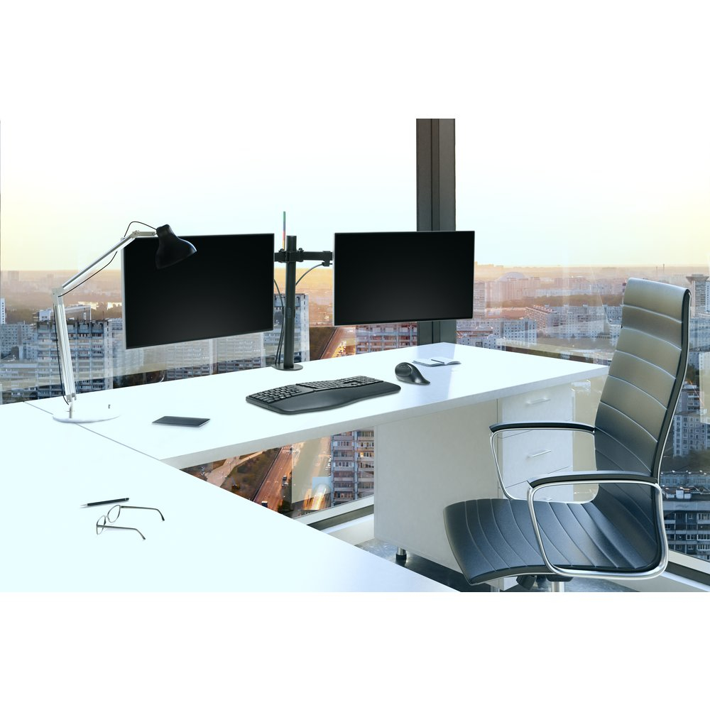 Modern bureau met dubbele monitor, toetsenbord, muis en bureaulamp, uitkijkend op stadsgezicht.