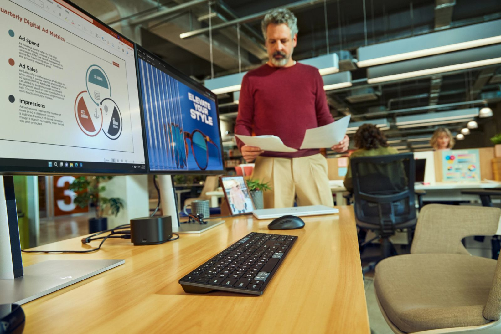Uomo in ufficio esamina carte vicino a monitor duali che mostrano metriche pubblicitarie e occhiali eleganti.