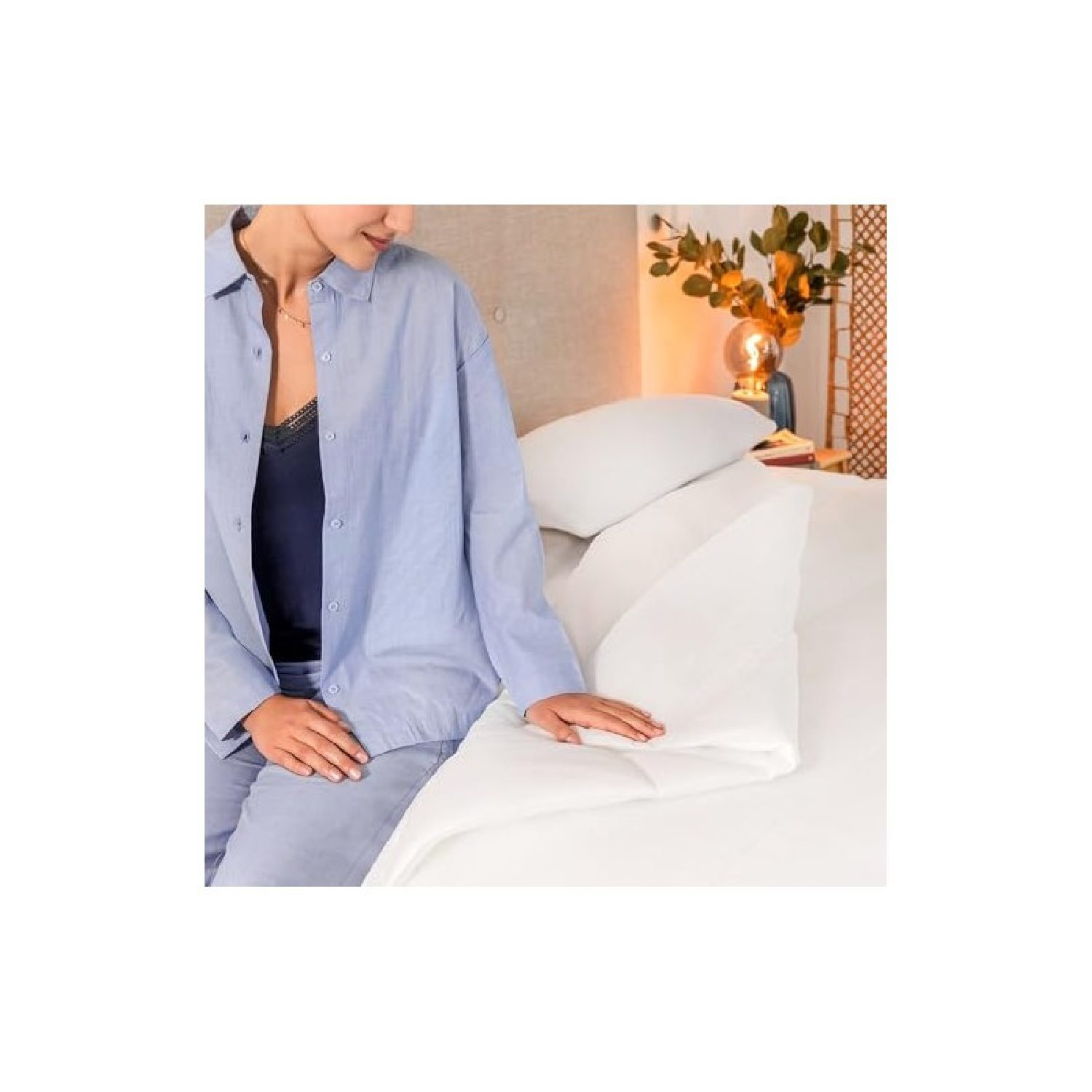 Una donna in pigiama blu è seduta su un letto bianco vicino a un cuscino a cuneo.