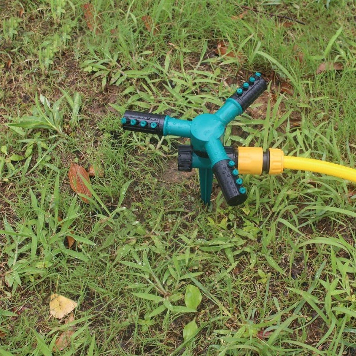Un irrigatore da giardino verde e nero a tre bracci con tubo giallo collegato, sull'erba.