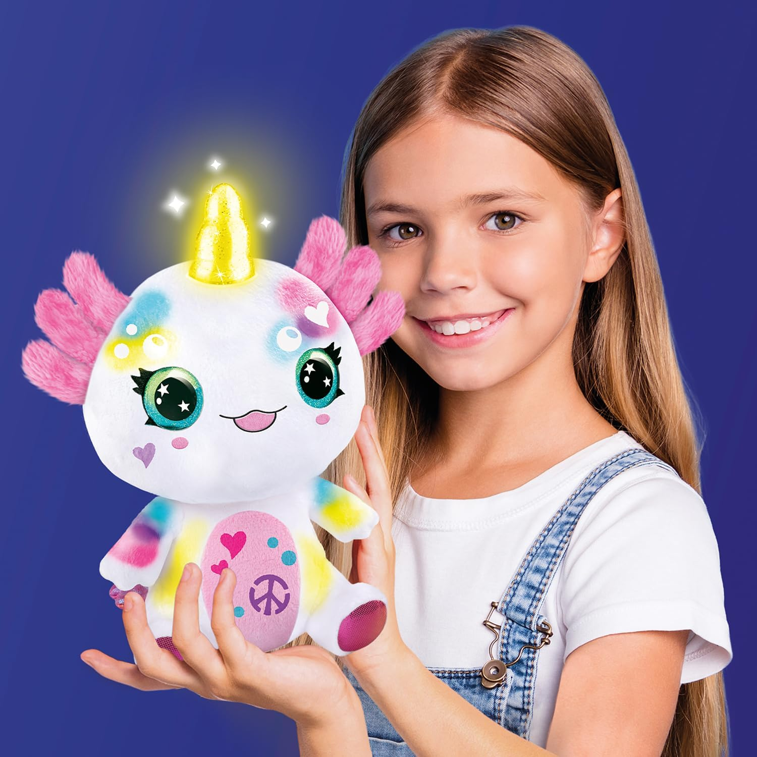 Una bambina sorridente tiene un peluche bianco arcobaleno con pinne rosa e corno giallo.