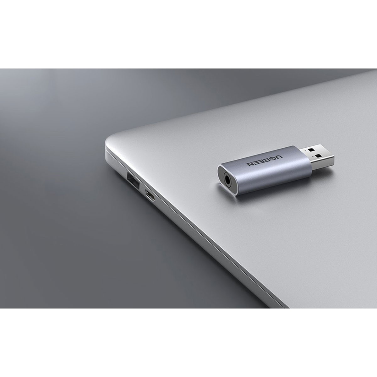 Srebrny adapter audio USB z gniazdem 3,5 mm leży na srebrnym laptopie.
