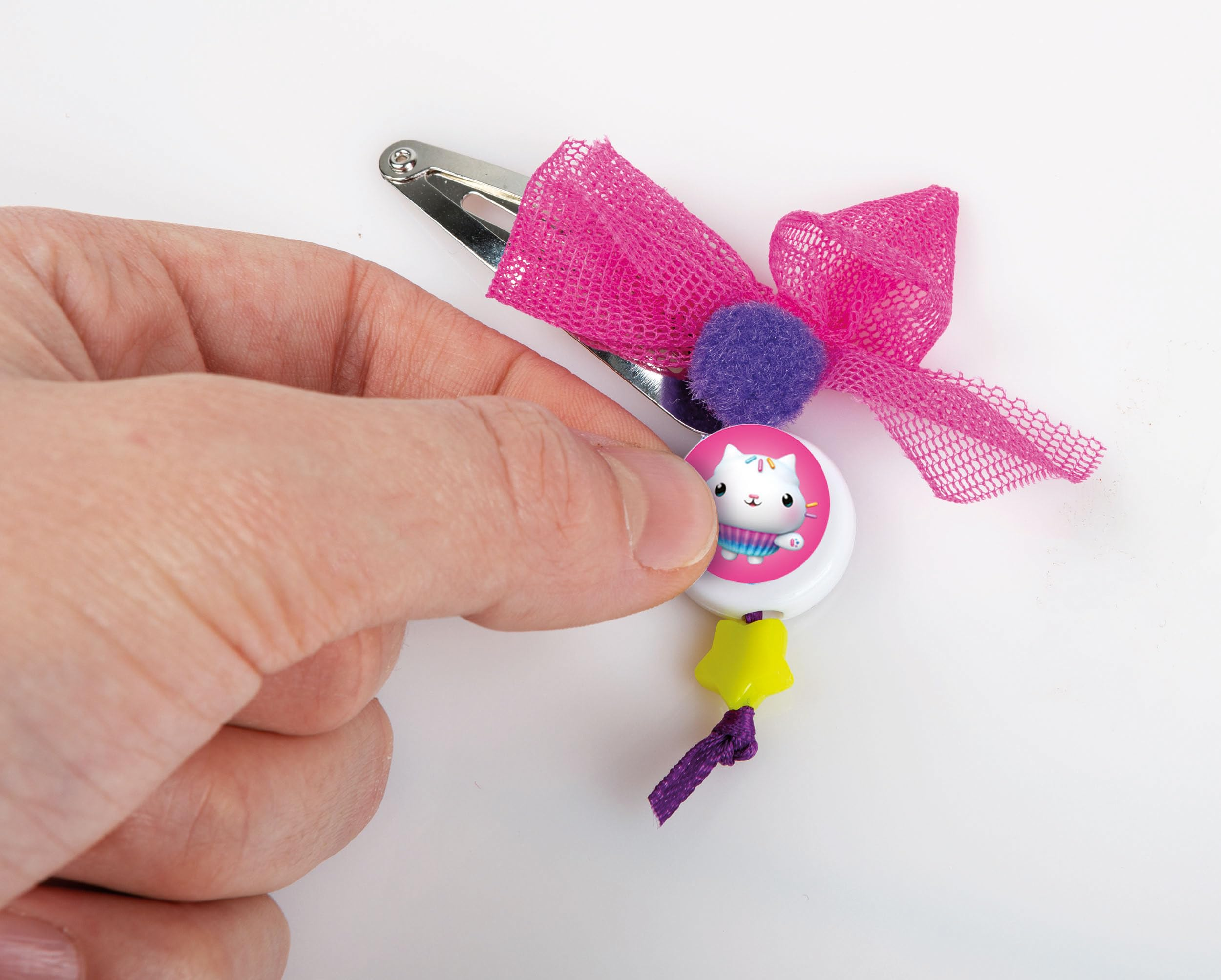 Una mano tiene un fiocco rosa con un pompon viola, un ciondolo di gatto e una stella gialla.