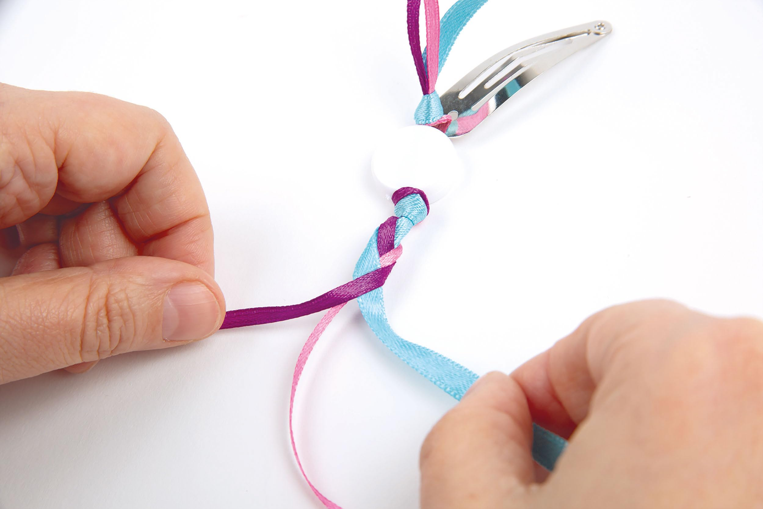 Mani intrecciano nastri rosa, viola e blu attorno a una perlina bianca e una clip d'argento.