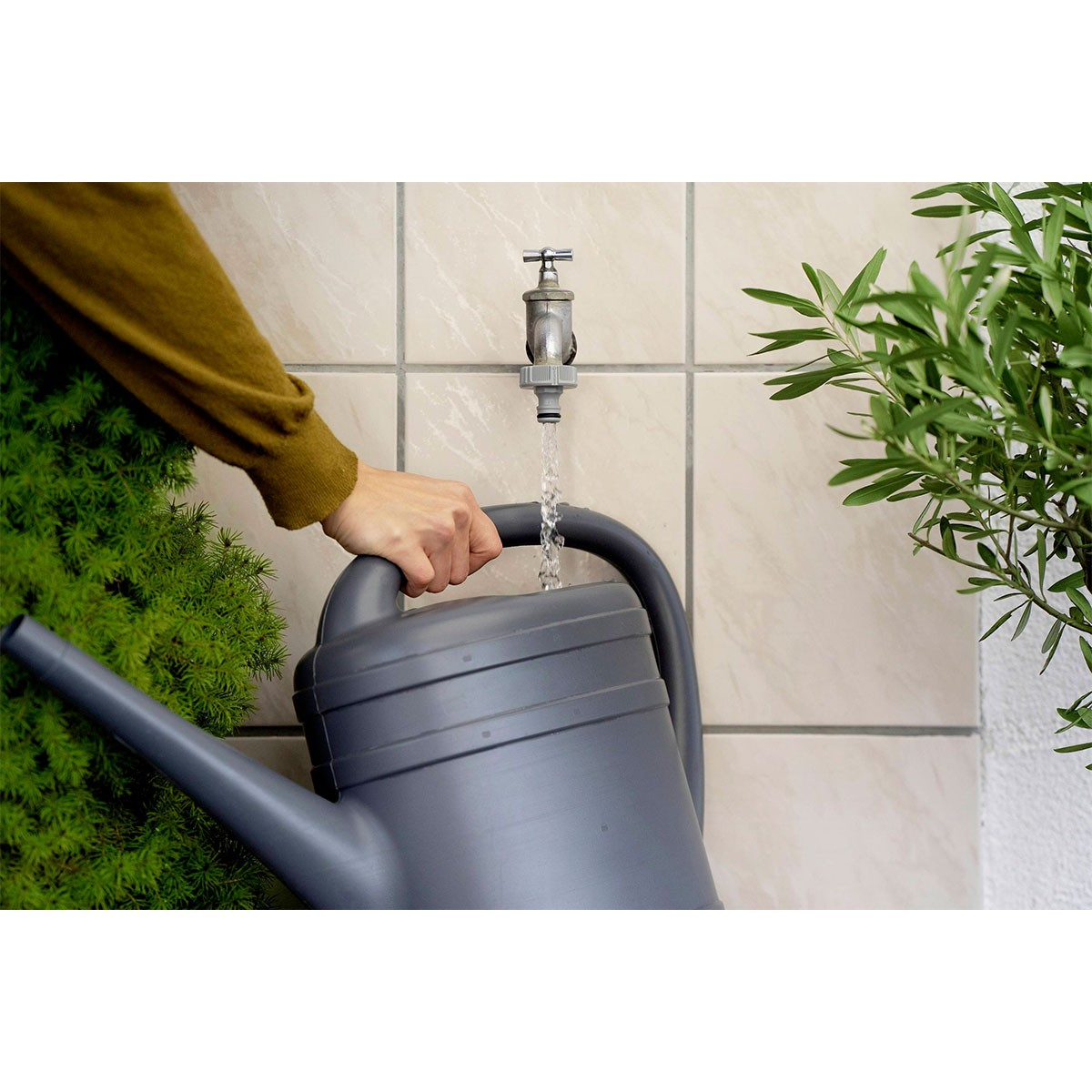 Una mano riempie un annaffiatoio grigio da un rubinetto da giardino contro un muro piastrellato.