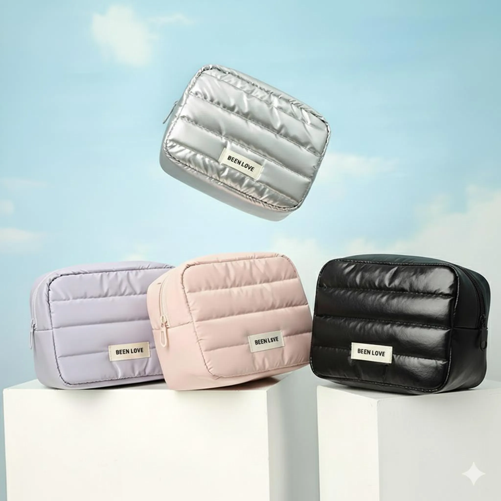 Vier gesteppte Kosmetiktaschen in Silber, Lila, Rosa und Schwarz schweben vor blauem Himmel.