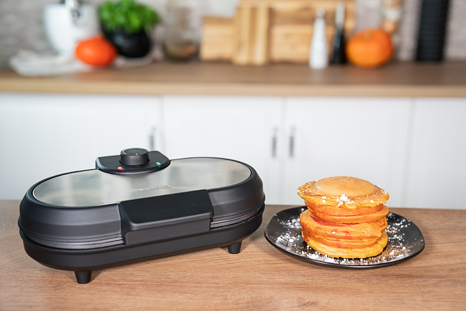 Ein schwarzer Waffelautomat steht neben einem Stapel goldbrauner Pfannkuchen mit Puderzucker.