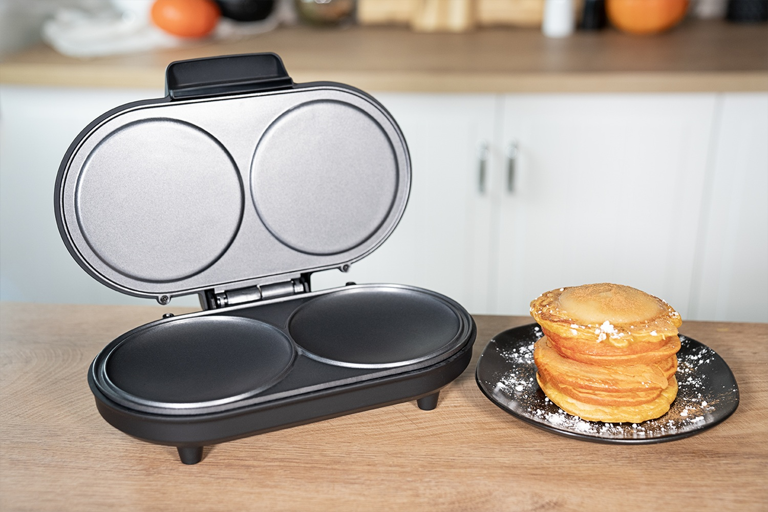 Offener elektrischer Pfannkuchengrill mit zwei runden Platten, neben einem Pfannkuchenstapel.