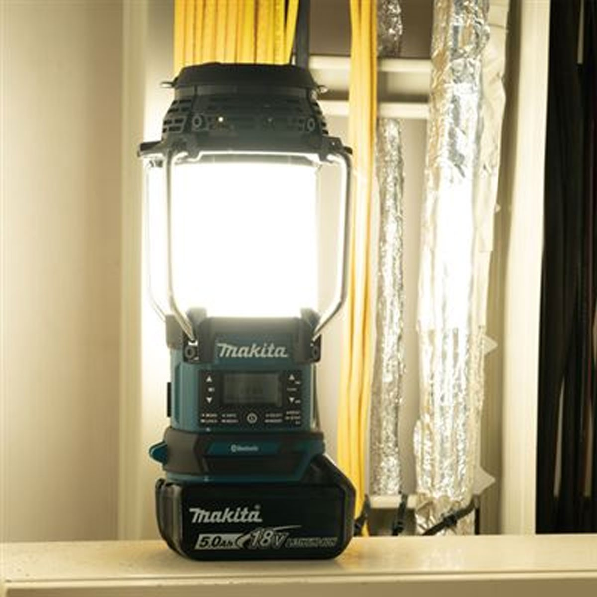 Une lanterne Makita lumineuse au corps turquoise et noir est sur une étagère.