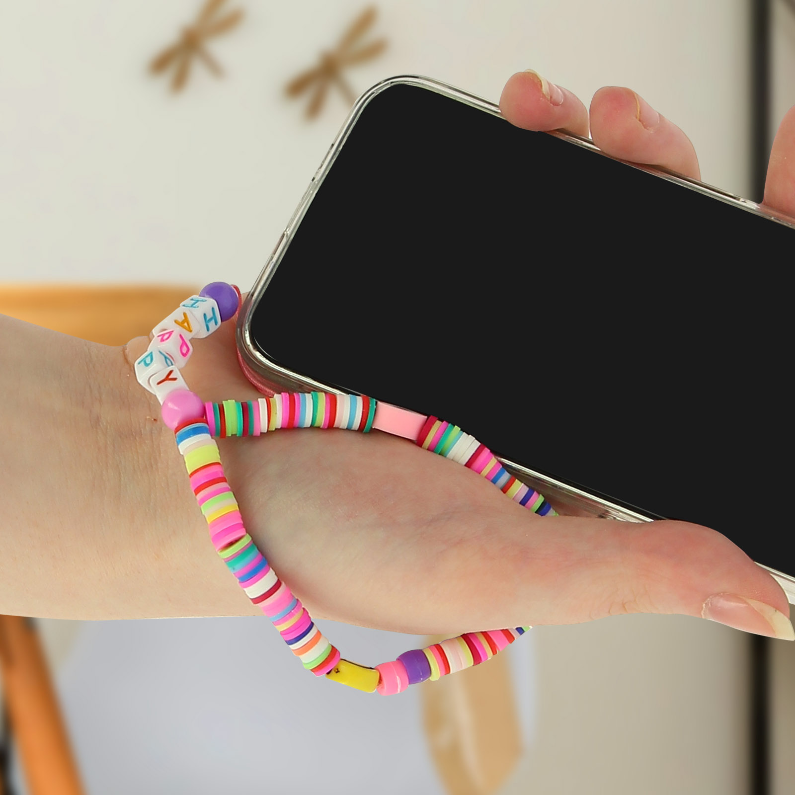 Una mano tiene un telefono con un cinturino colorato di perline che dice 'HAPPY'.
