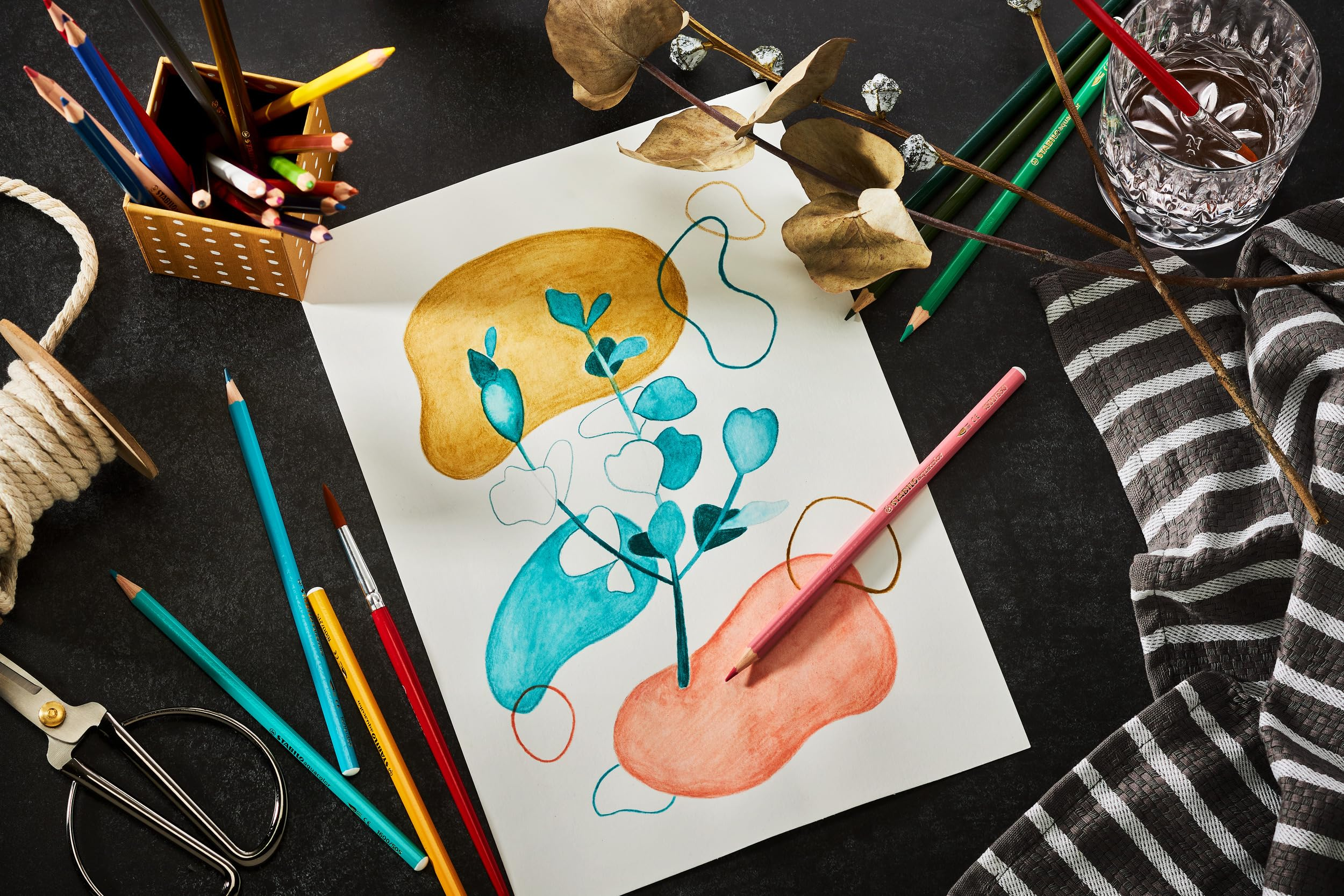 Materiali artistici su superficie scura: disegno, matite, corda, forbici, stoffa e foglie secche.