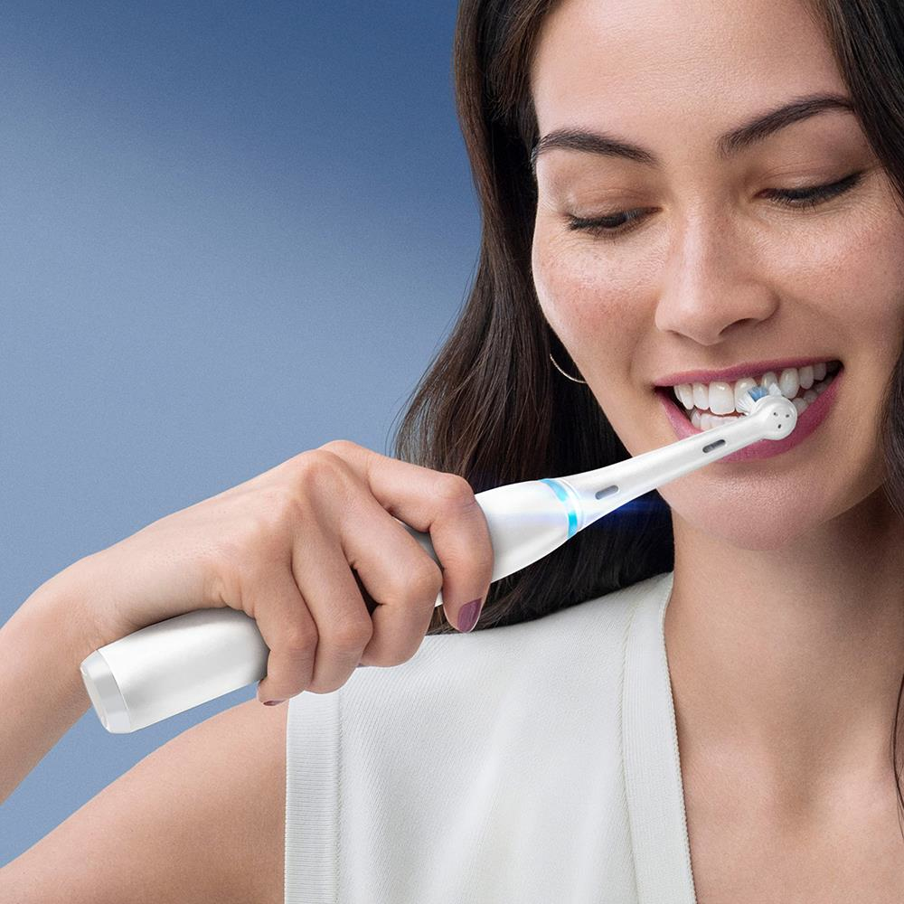 Una donna dai capelli scuri si lava i denti con uno spazzolino elettrico bianco su sfondo blu.