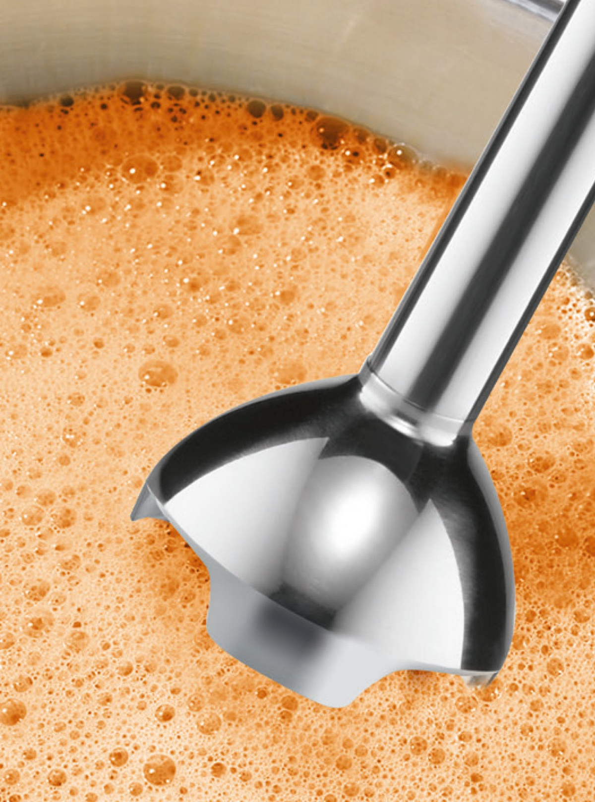 Nahaufnahme eines silbernen Stabmixer-Kopfs in schaumiger oranger Flüssigkeit.