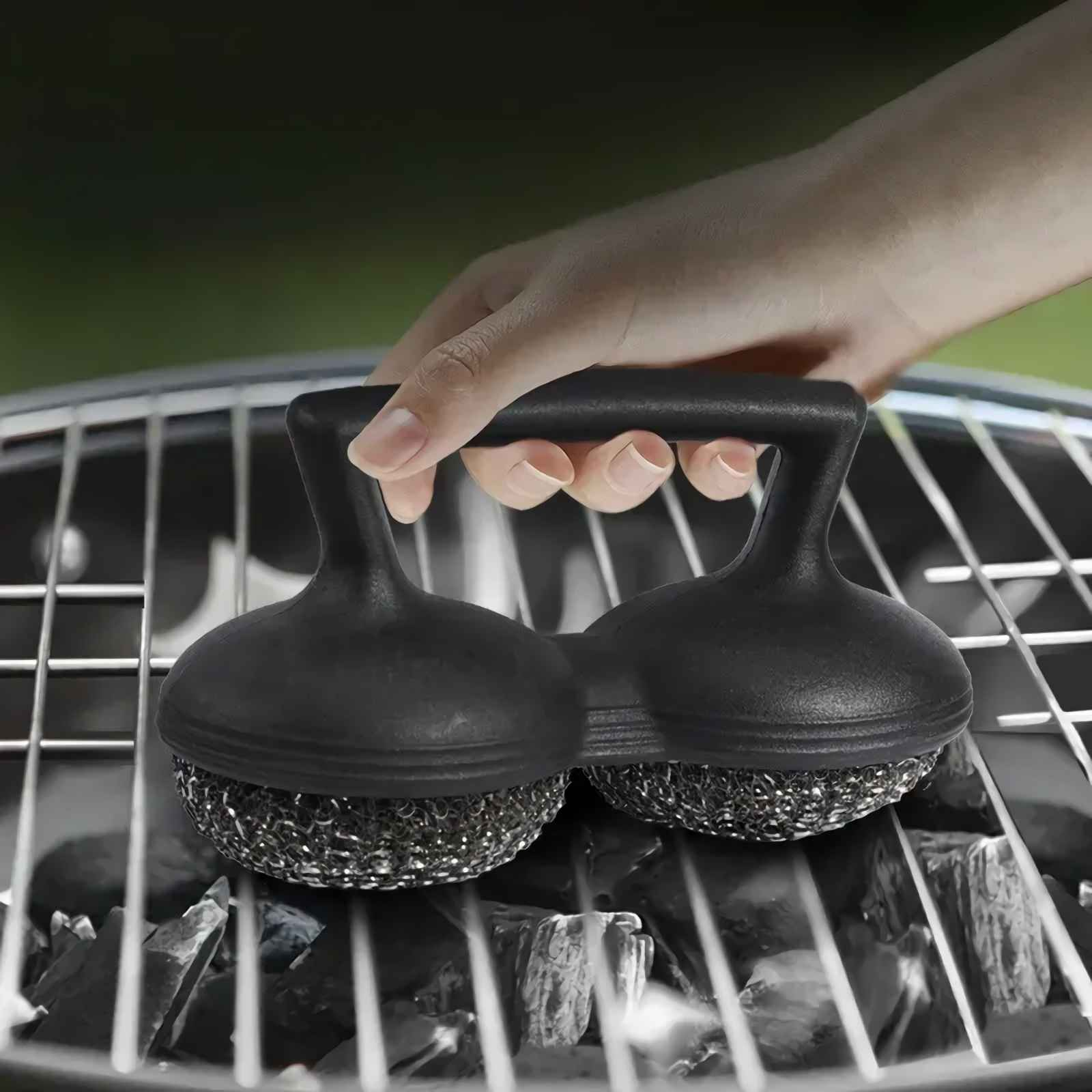 Hand hält eine schwarze Grillbürste mit zwei Bürsten über einem Holzkohlegrill.
