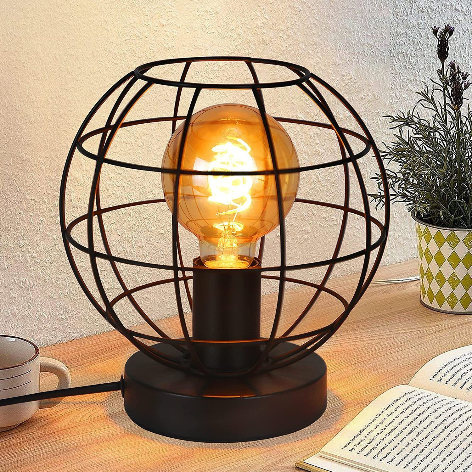 Una lampada a gabbia in metallo nero con lampadina visibile è su una scrivania di legno accanto a un libro e una tazza.