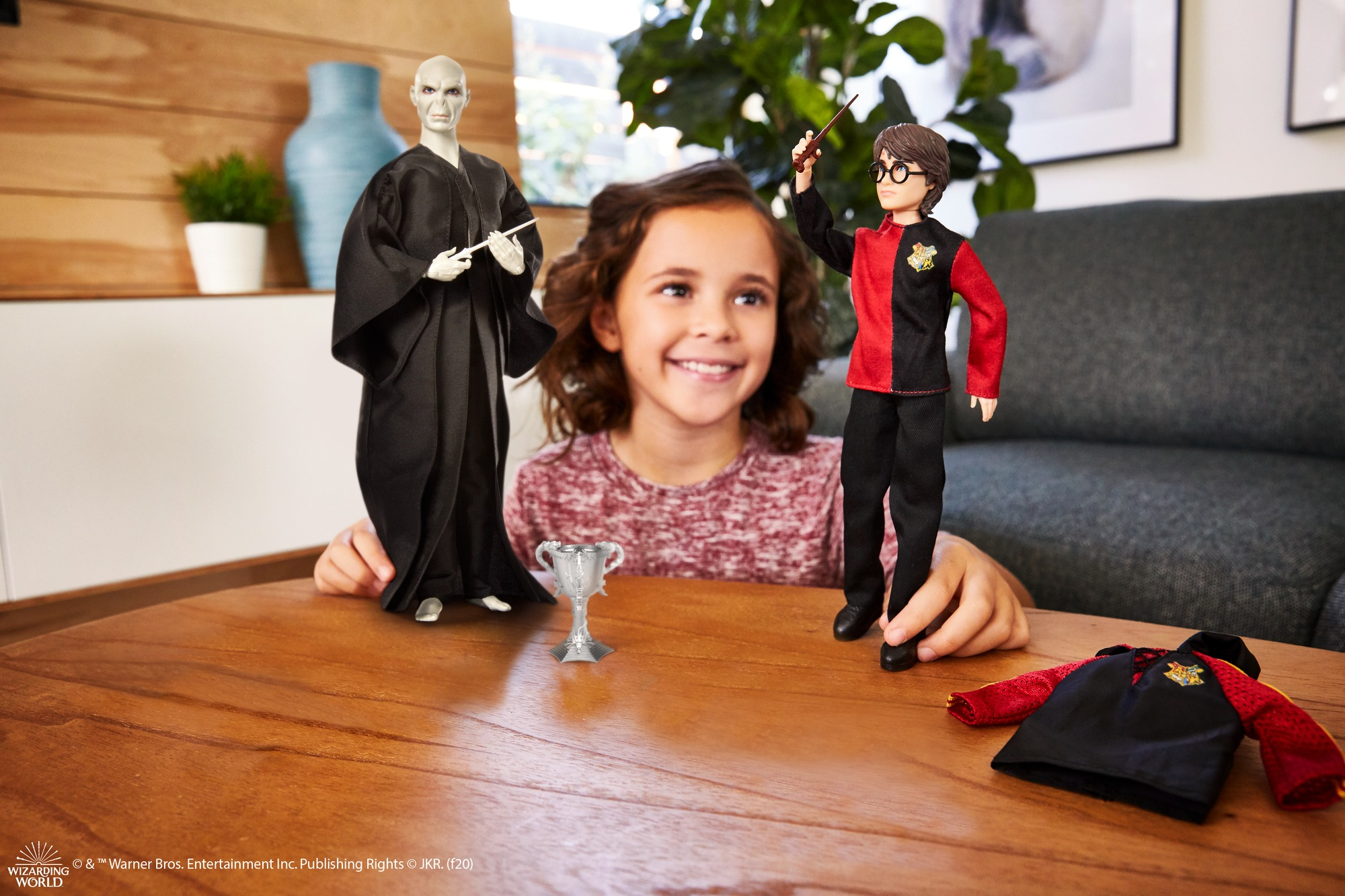 Bambole di Voldemort e Harry Potter su un tavolo con una bambina dietro. Un trofeo davanti.