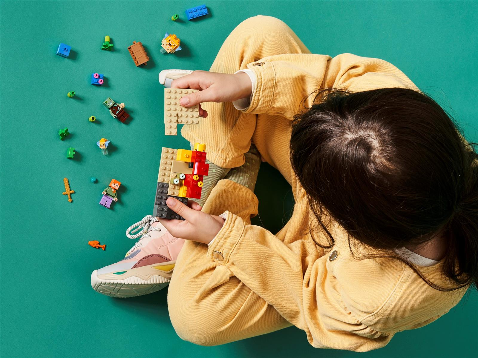 Ein Kind in gelber Kleidung baut mit bunten Legosteinen auf einem türkisfarbenen Hintergrund.