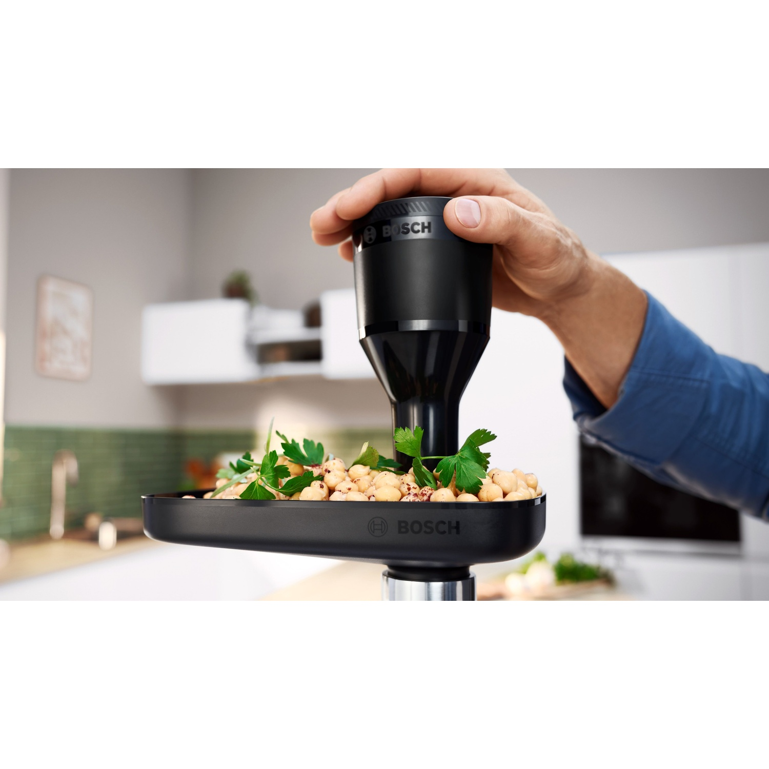 Una mano tiene un accessorio nero Bosch per robot da cucina sopra un vassoio di ceci e prezzemolo.