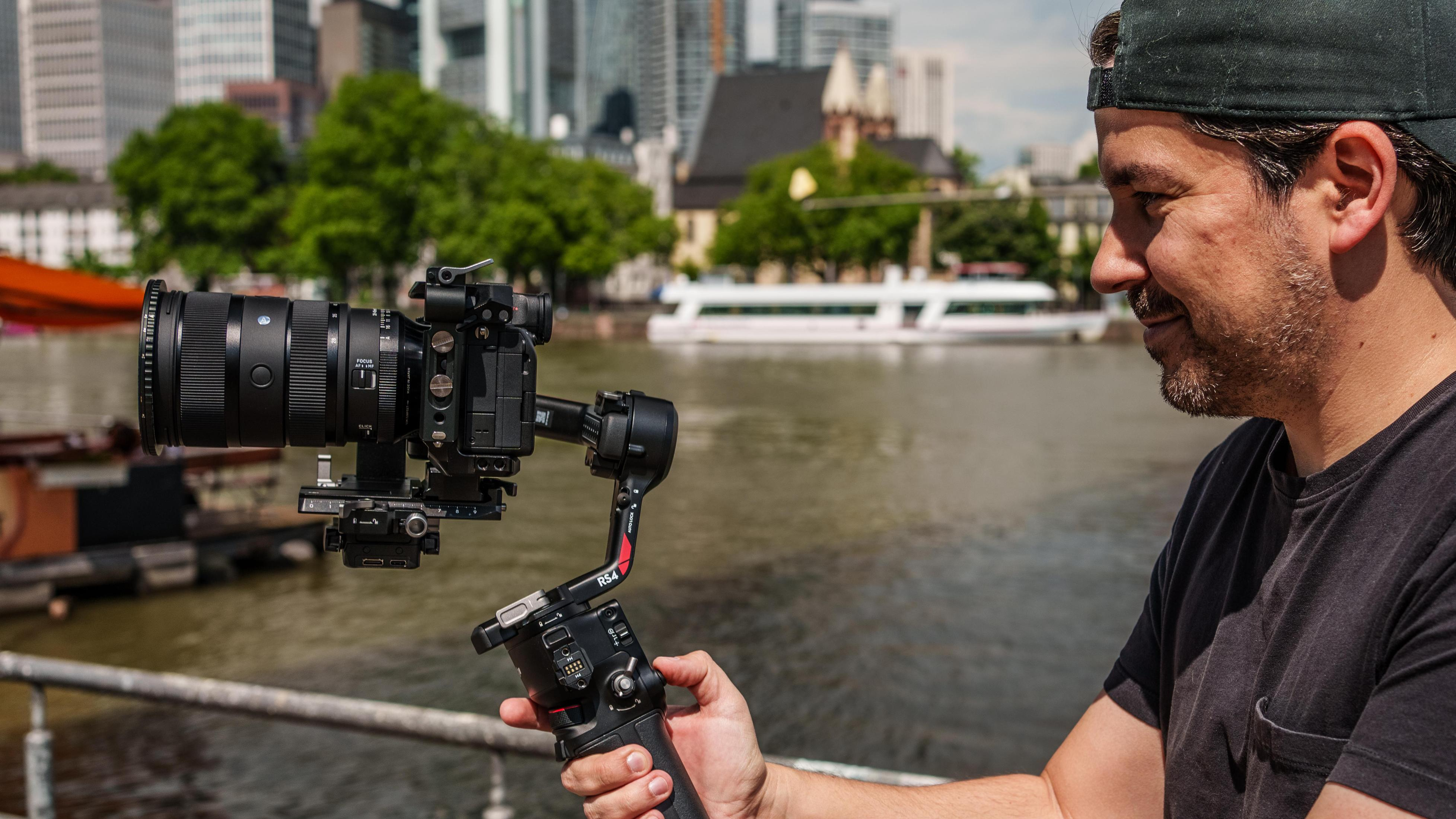 Mężczyzna trzyma gimbal z aparatem przed rzeką i panoramą miasta.