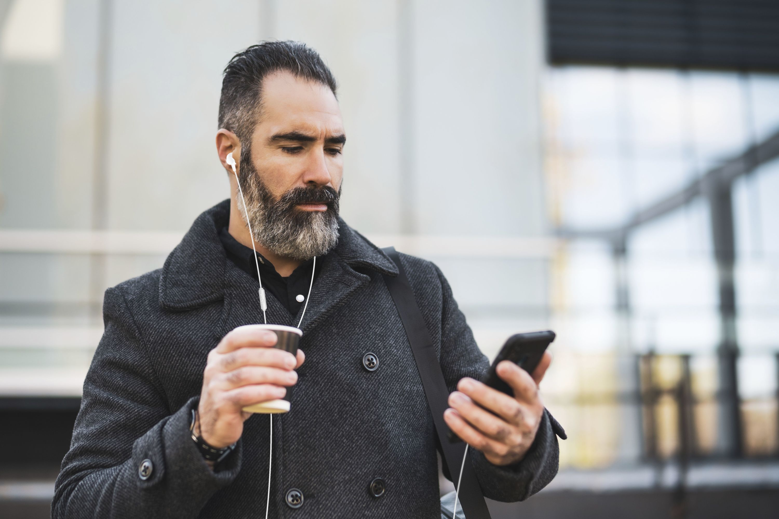 A man with a beard listens to music with earphones, holding a coffee cup.