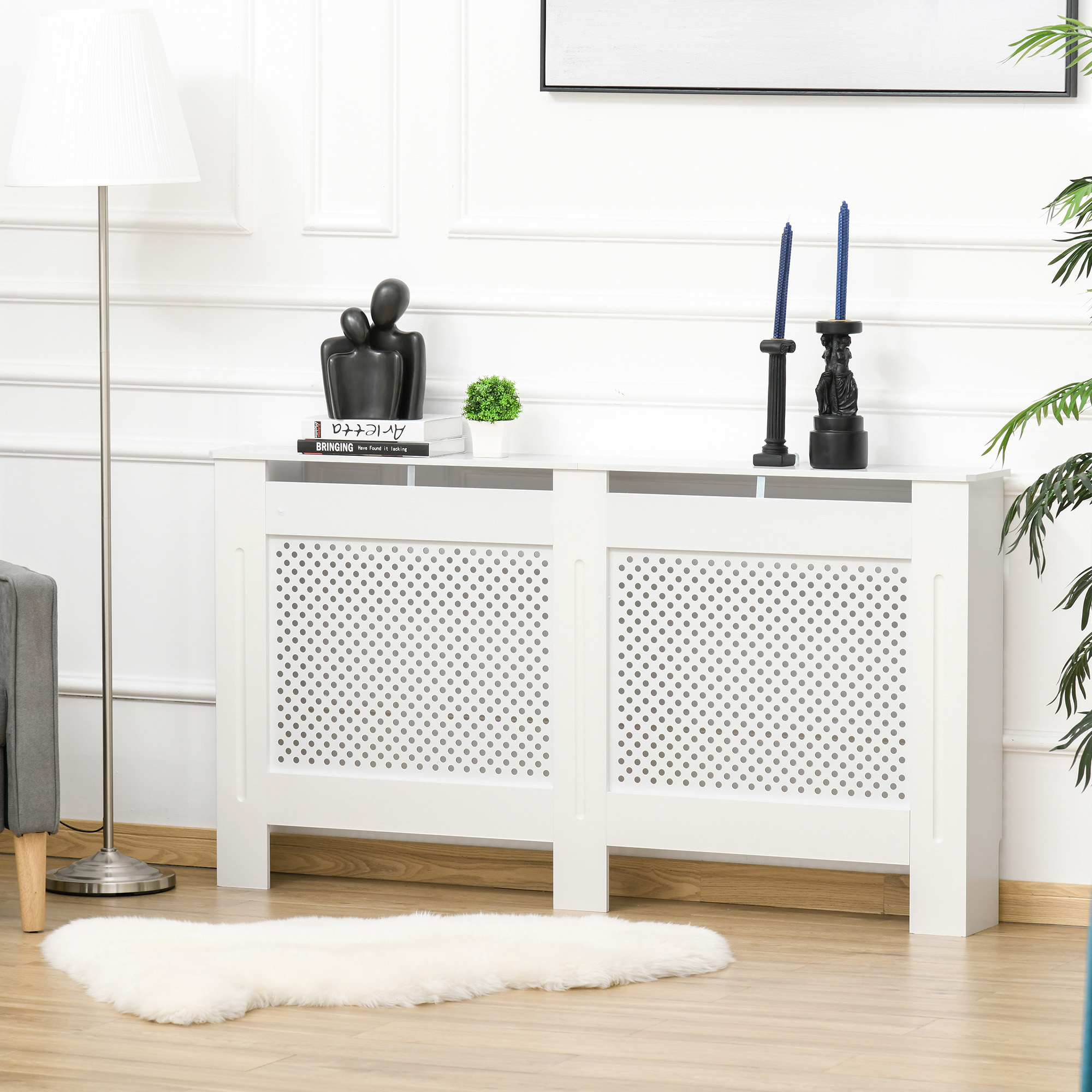 Copritermosifone bianco con ante forate, su pavimento in legno. Lampada, decorazioni, libri e pianta.