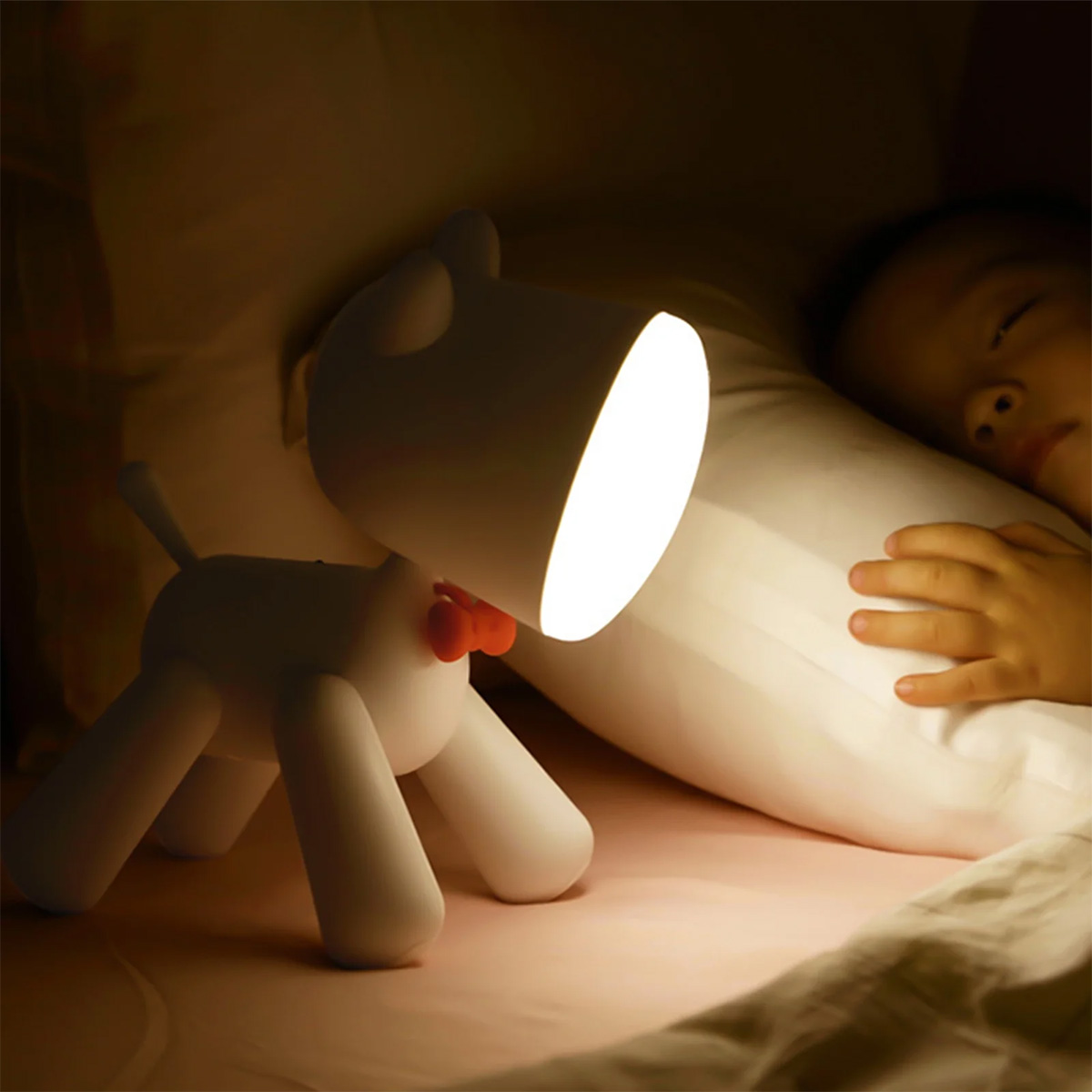 Eine weiße hundförmige Lampe mit roter Schleife leuchtet auf ein schlafendes Kind im Bett.