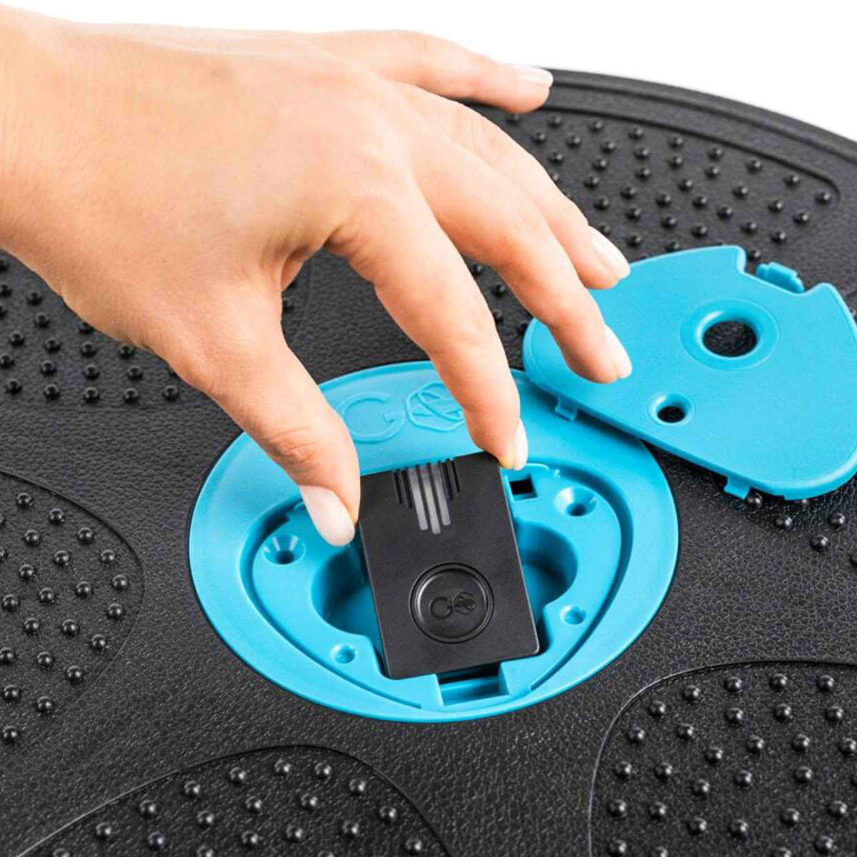 Eine Hand schiebt eine schwarze Batteriepackung in ein blaues Fach auf einer schwarzen, strukturierten Oberfläche.