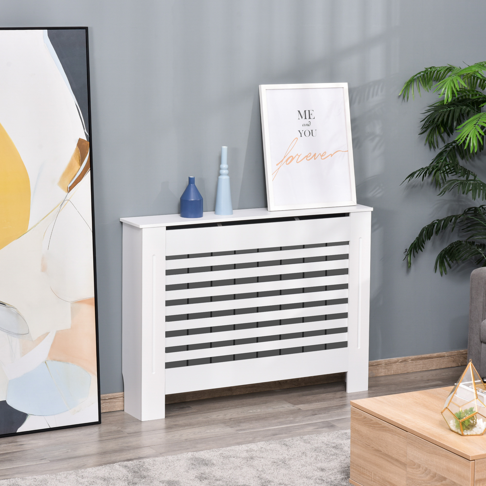 Una copertura per termosifone bianca con doghe orizzontali, due vasi e un'opera d'arte incorniciata su un muro grigio.