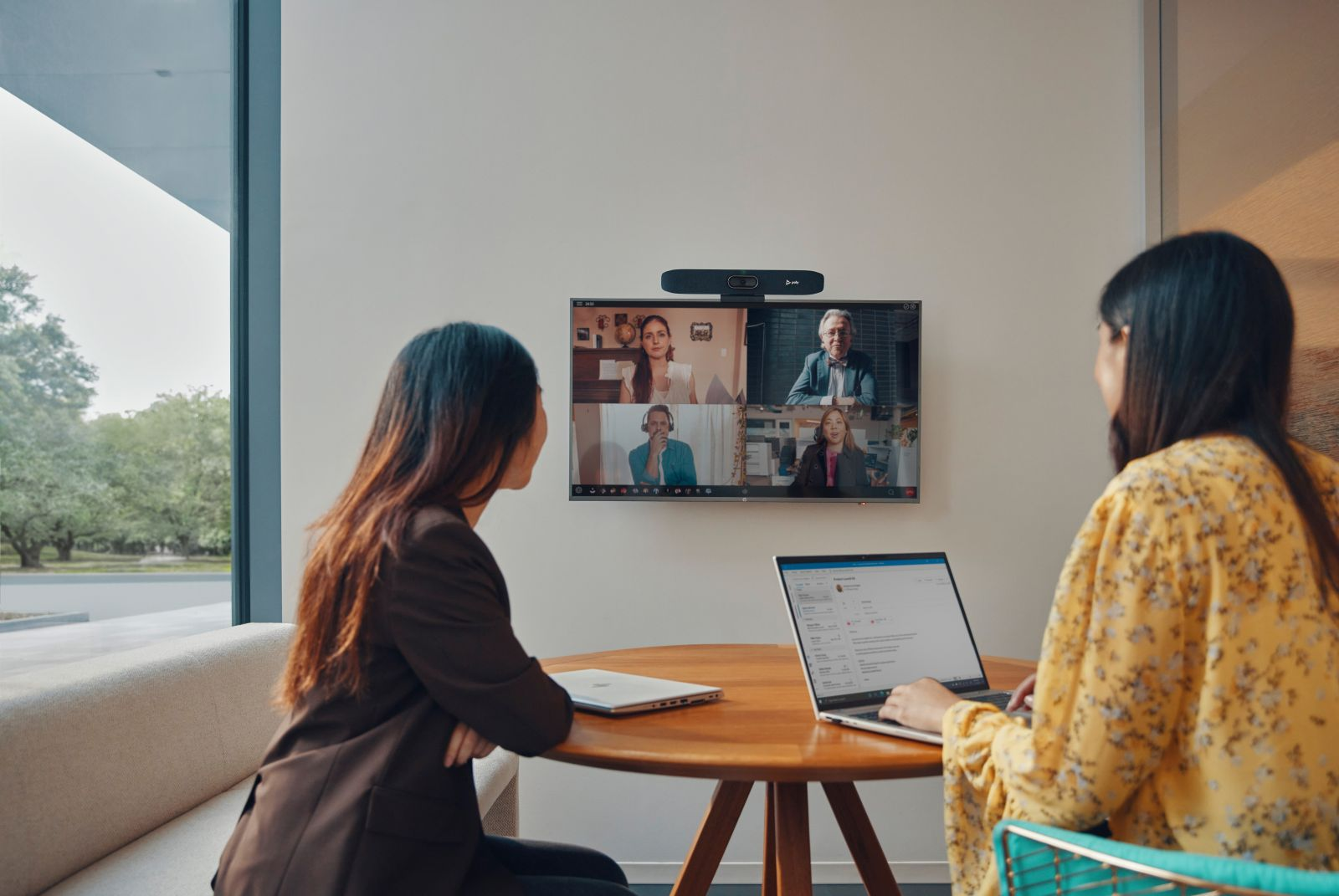 Dos mujeres en una oficina asisten a una videoconferencia con cuatro personas en pantalla. Un portátil está abierto.