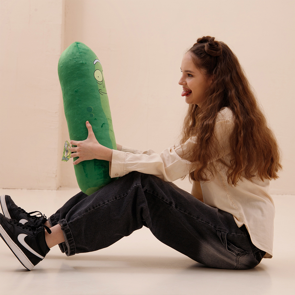 Ein Mädchen sitzt und hält ein grünes Pickle-Plüschtier, es streckt die Zunge heraus.