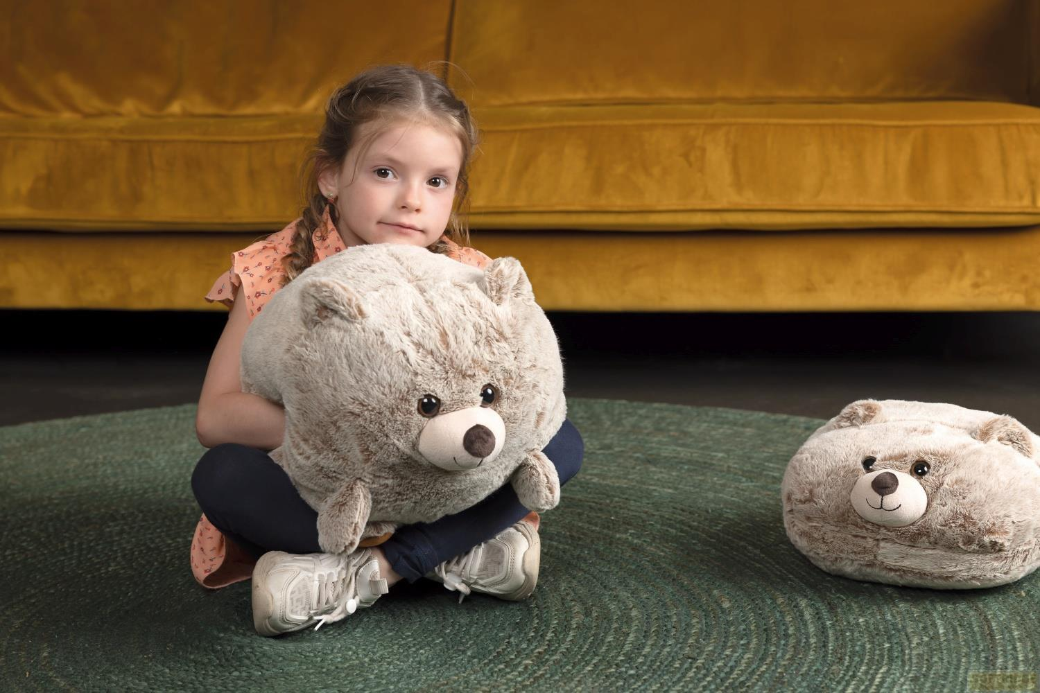 Ein Mädchen sitzt mit einem Teddybär auf einem Teppich vor einer gelben Couch.