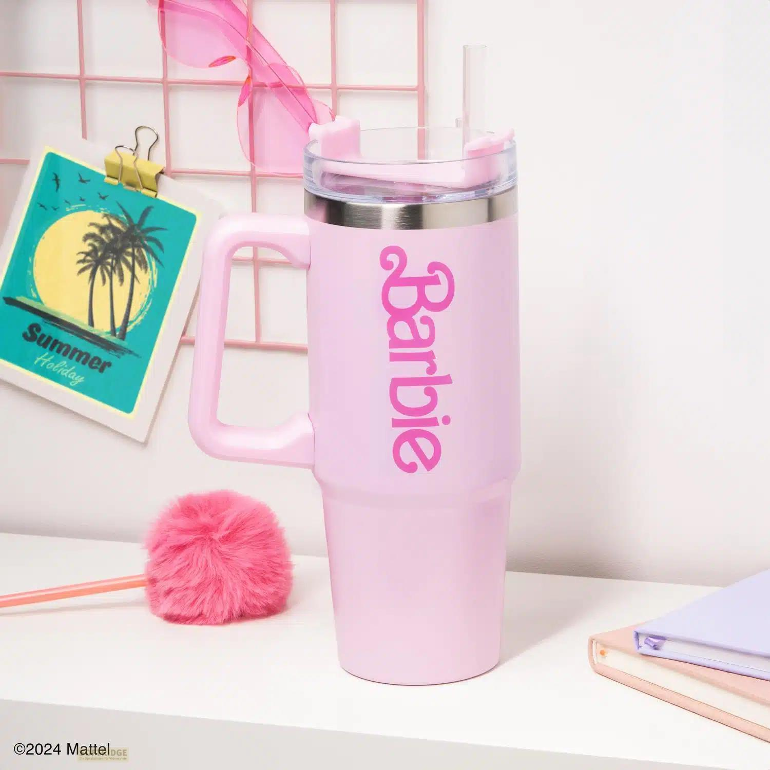 Un vaso Barbie rosa con pajita está sobre un escritorio blanco junto a una postal de vacaciones de verano y un pompón.