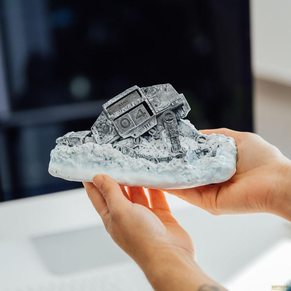 Ein metallisches AT-AT Walker-Modell auf einer schneebedeckten Basis, gehalten von zwei Händen. Der Kalender zeigt den 4. Mai.