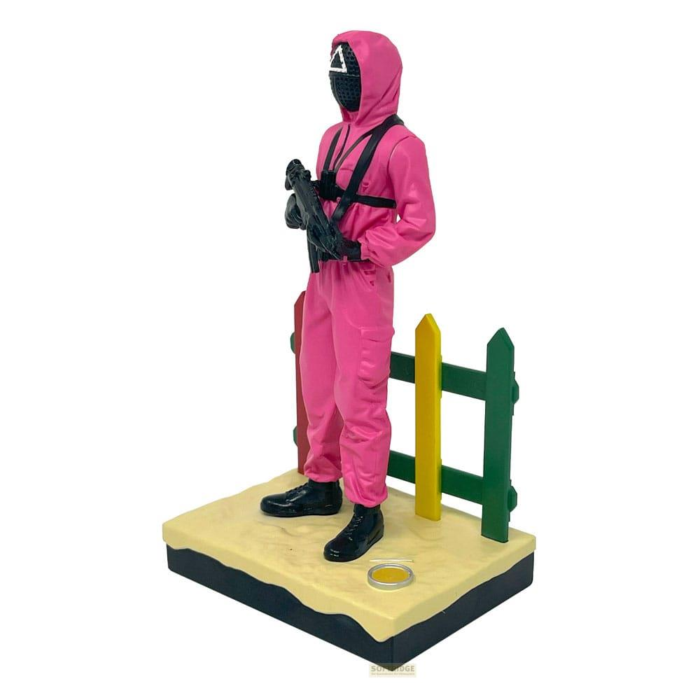 Figura de Squid Game con uniforme rosa y arma sobre arena y valla.