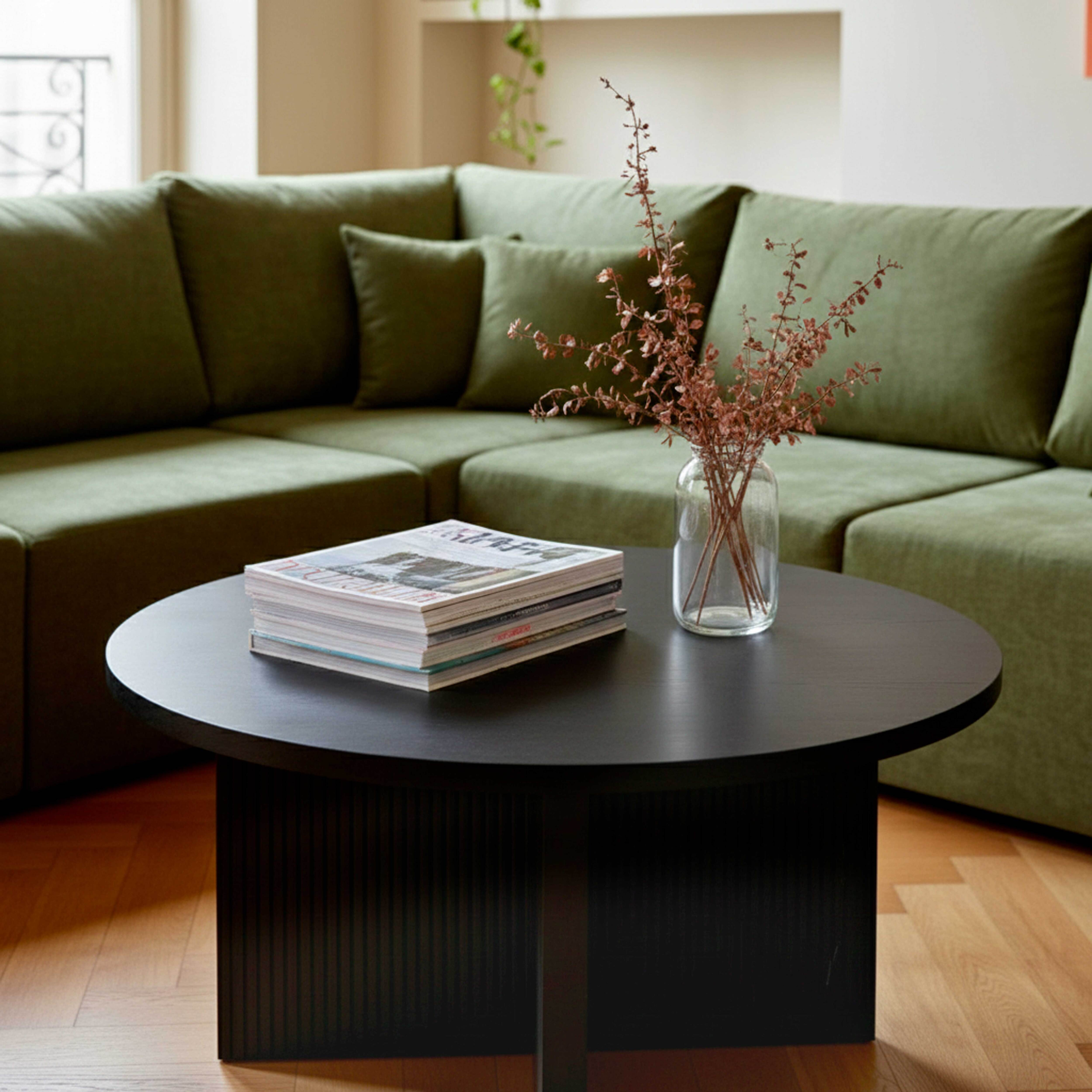 Ein runder schwarzer Couchtisch mit Zeitschriften und einer Vase mit Zweigen vor einem grünen Sofa.