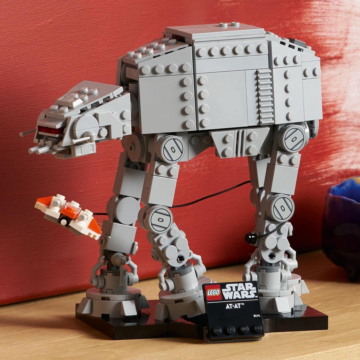Kırmızı arka planlı gri Lego AT-AT modeli ve küçük turuncu araç.
