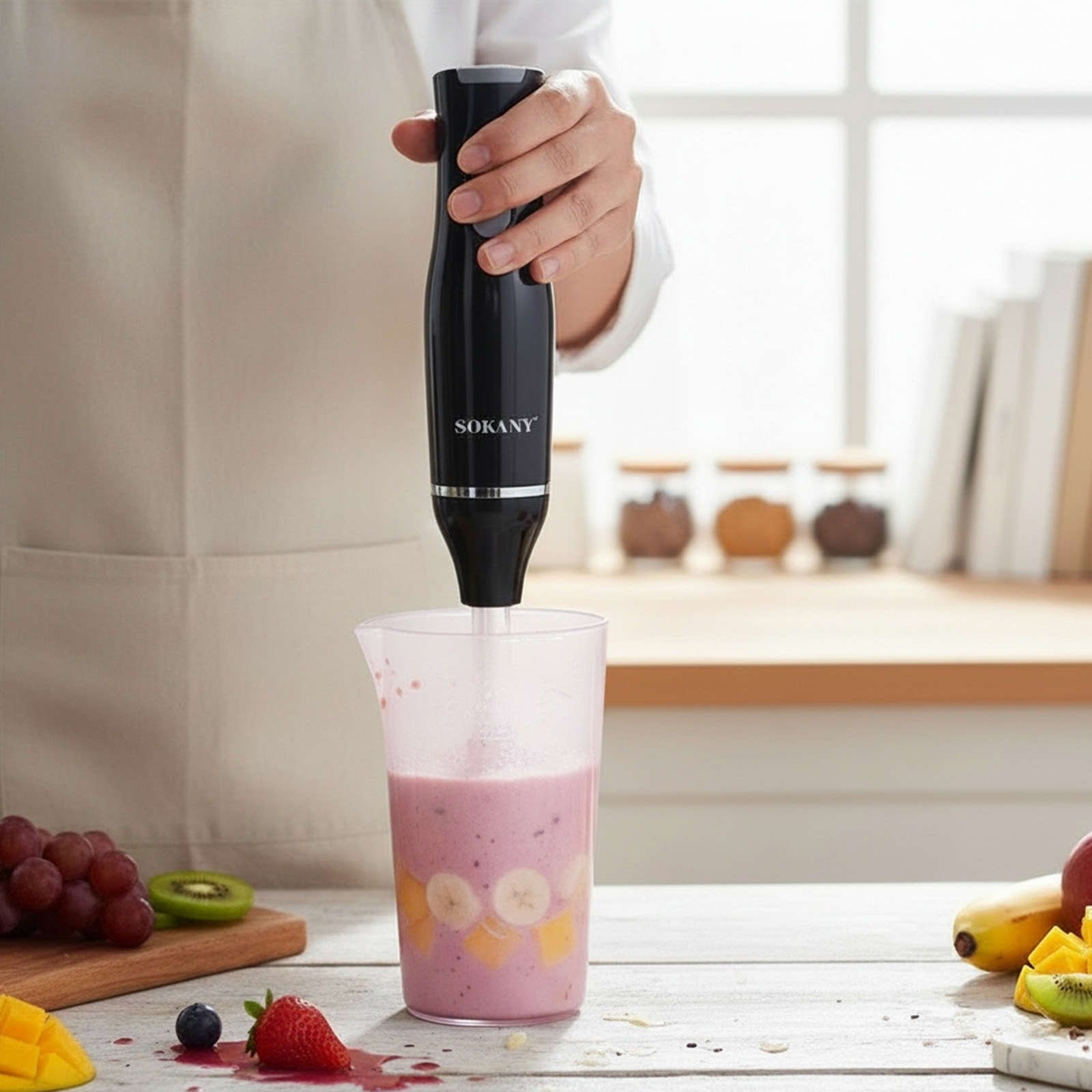 Eine Hand hält einen schwarzen Stabmixer über einem Glas Smoothie mit Obst.