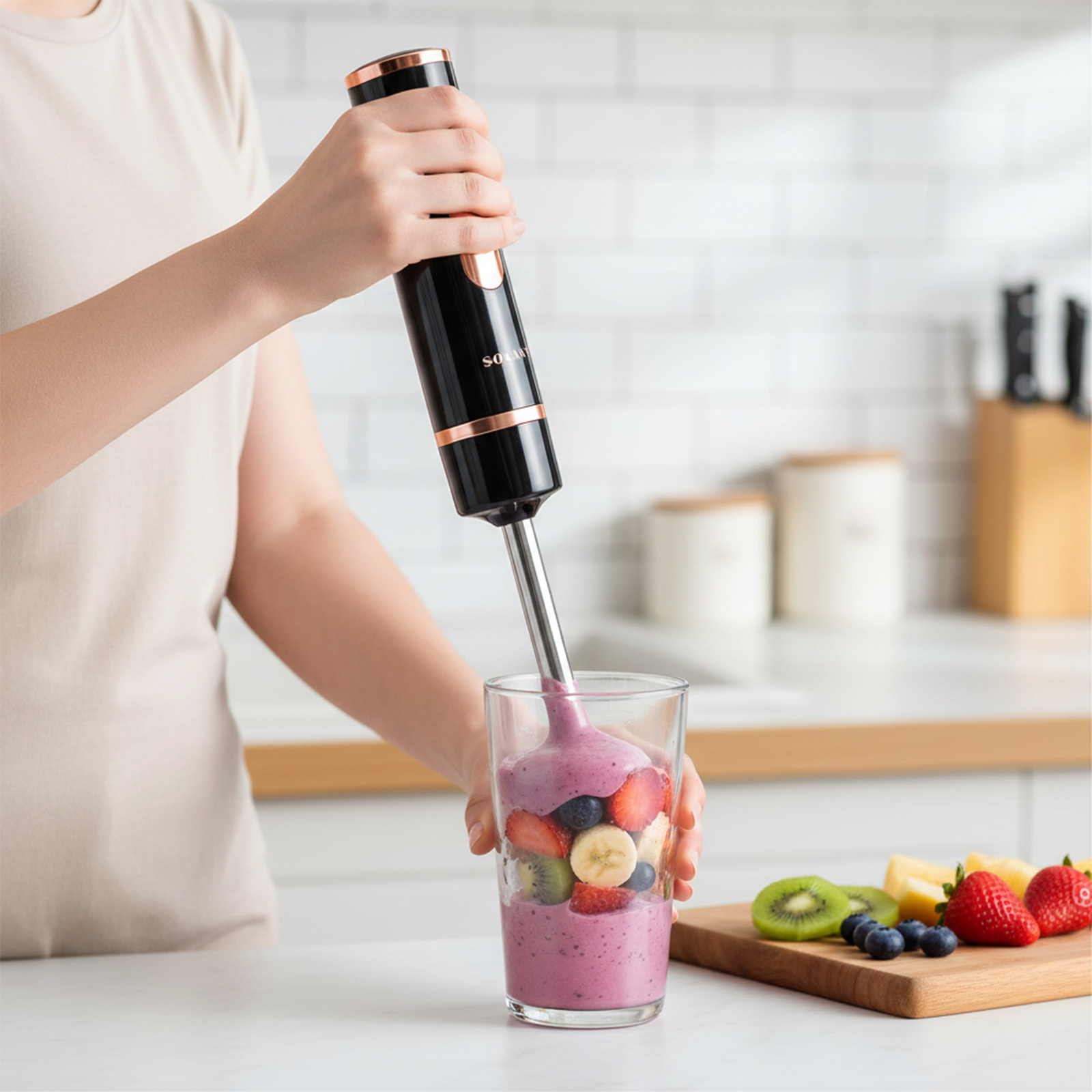 Eine Hand hält einen schwarzen Stabmixer über einem Glas mit Fruchtstücke.