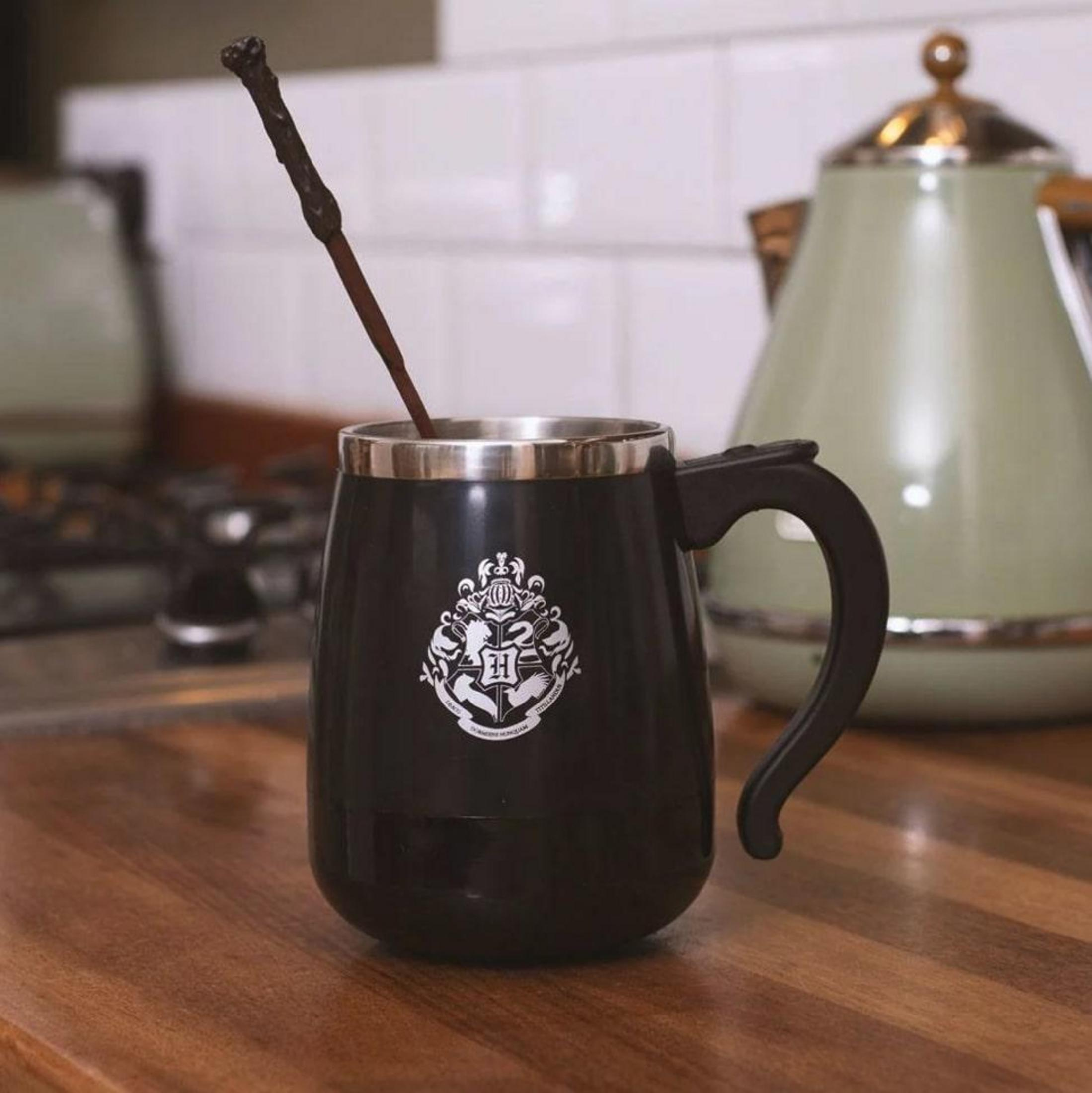 Tasse noire avec blason de Poudlard et touilleur baguette, près d'une bouilloire verte.