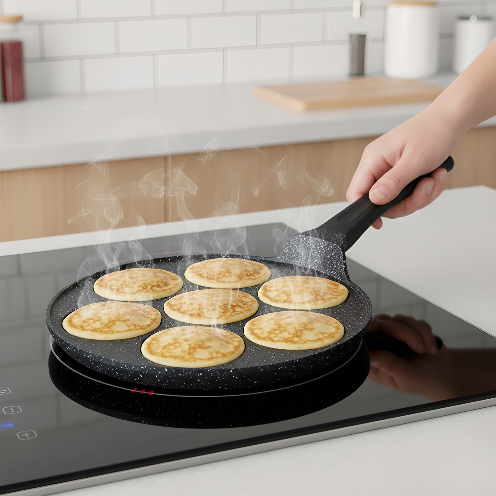Une main tient une poêle avec sept pancakes dorés cuisant sur une plaque de cuisson noire.