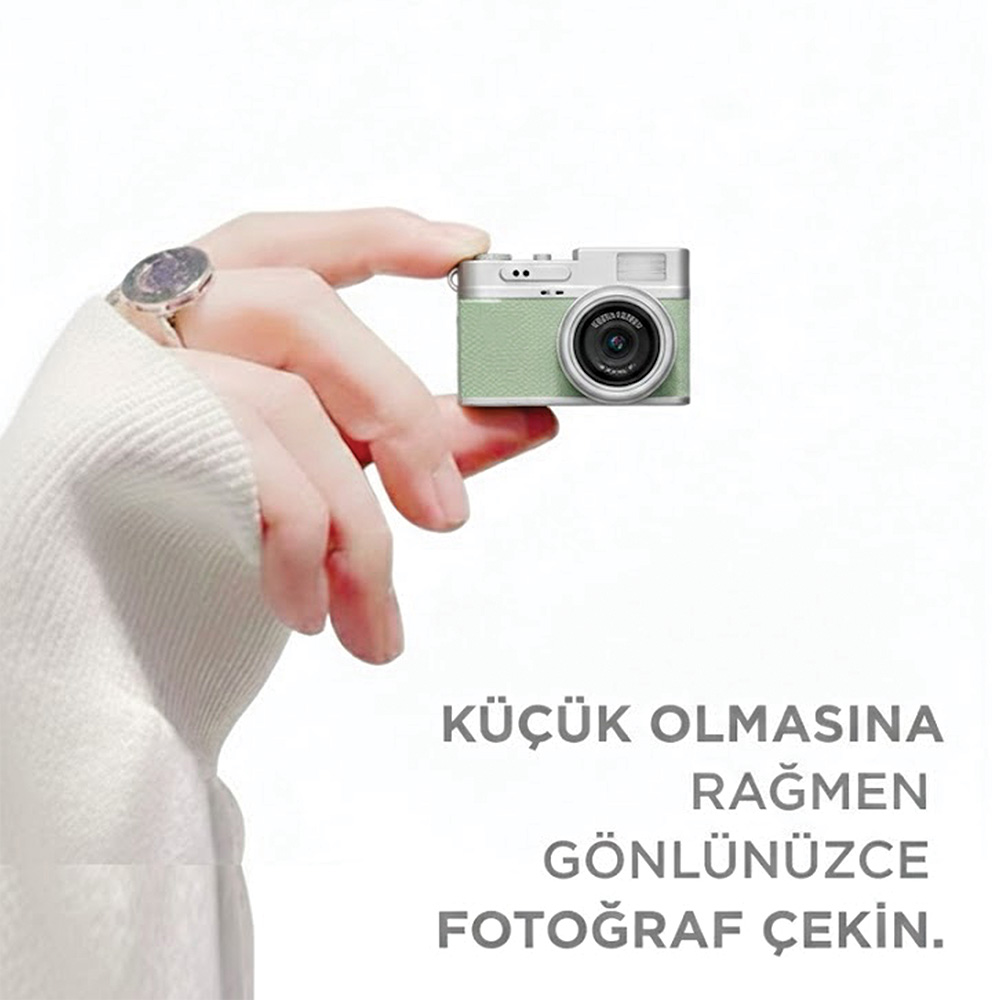 Bir el, minik yeşil bir fotoğraf makinesi tutuyor. Elde yüzük ve kazak var.