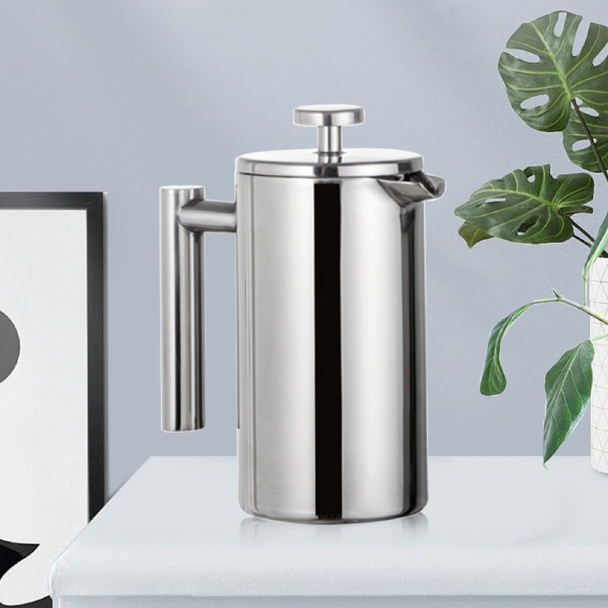 Elegancka, srebrna prasa francuska stoi na białej powierzchni obok ramki i rośliny.