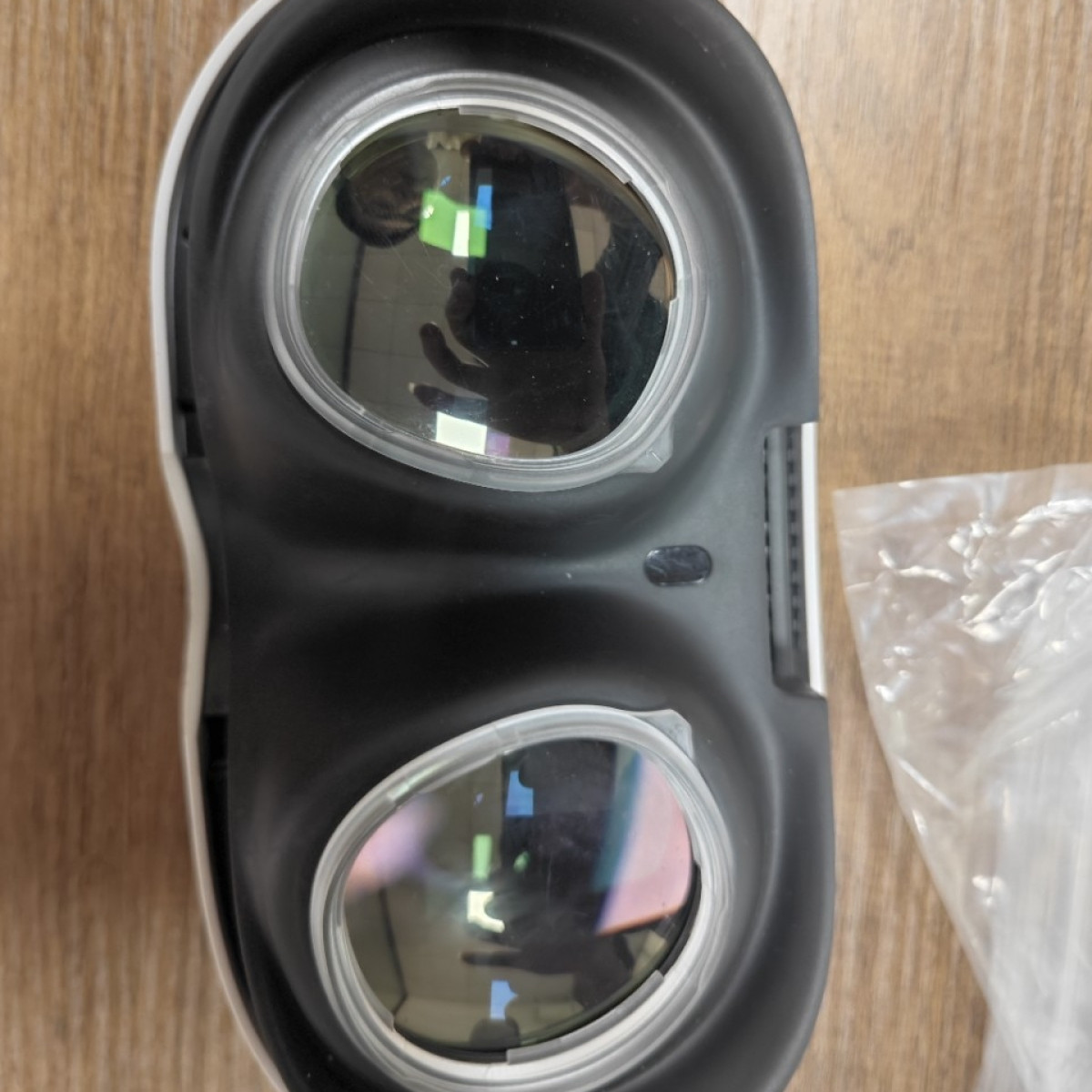 Czarny zestaw VR z dużymi soczewkami leży na drewnianej powierzchni. Po prawej stronie znajduje się przezroczysta torba plastikowa.