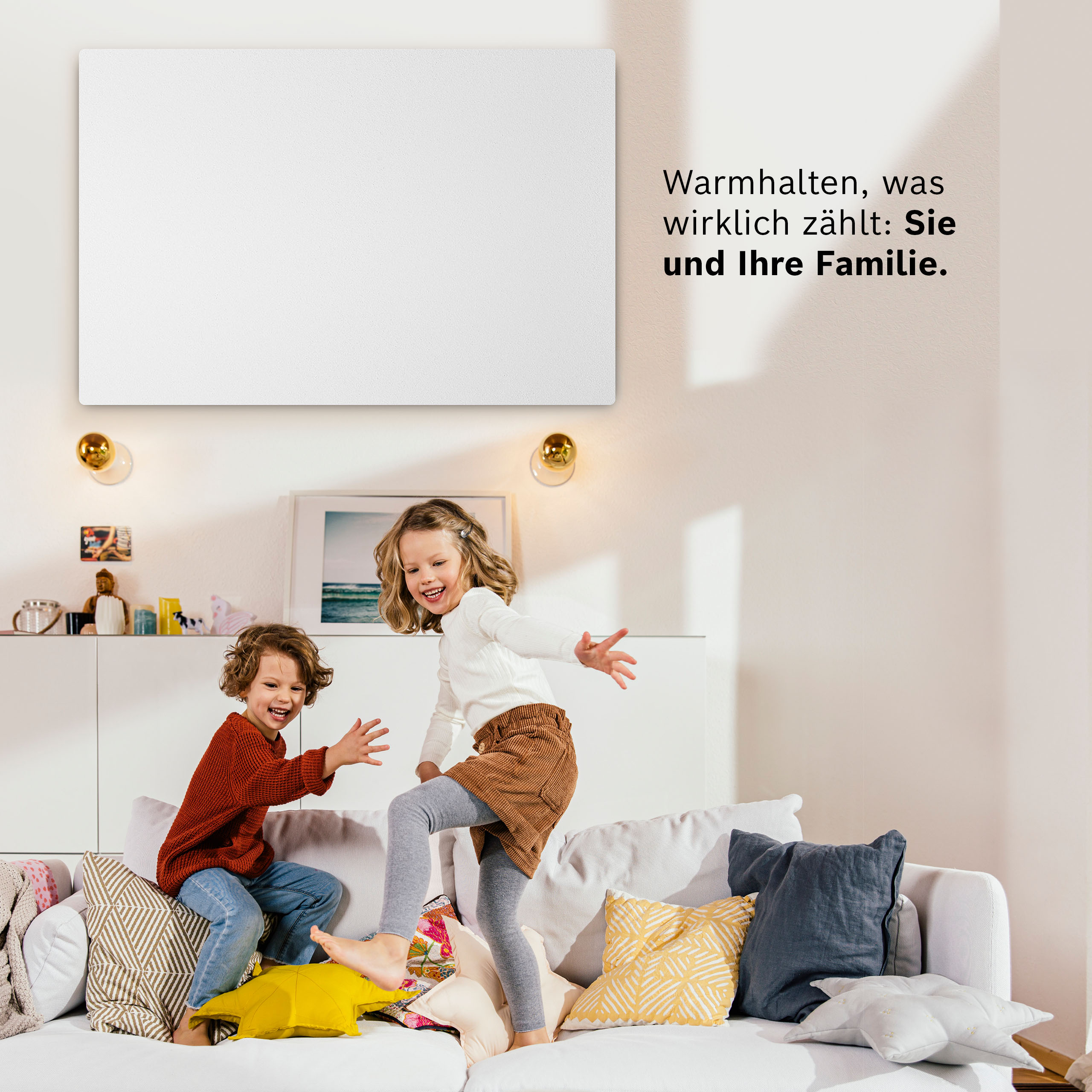 Zwei lächelnde Kinder springen auf ein Sofa mit bunten Kissen. Eine weiße Leinwand hängt darüber.