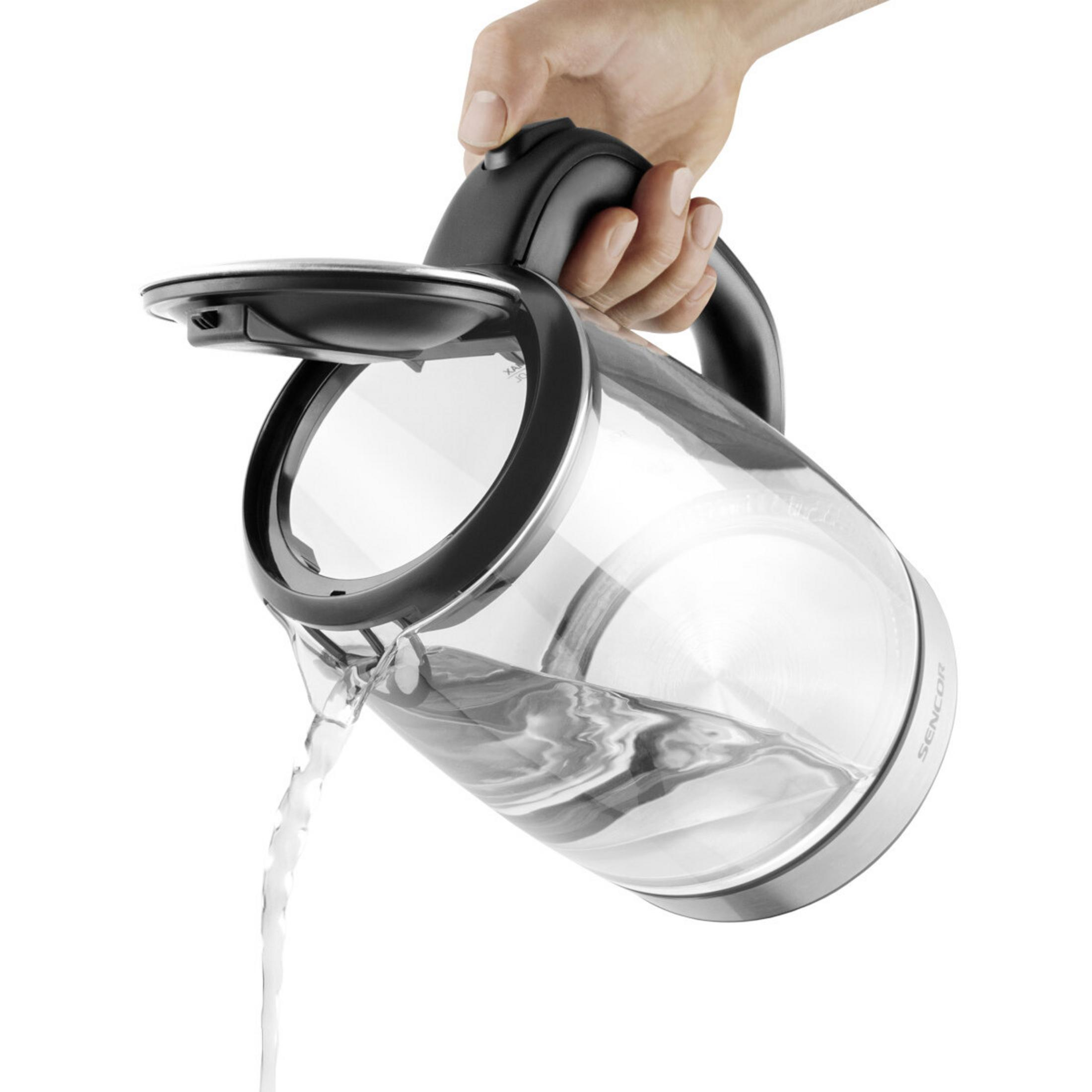 Een hand giet water uit een glazen waterkoker met een zwart deksel en handvat.