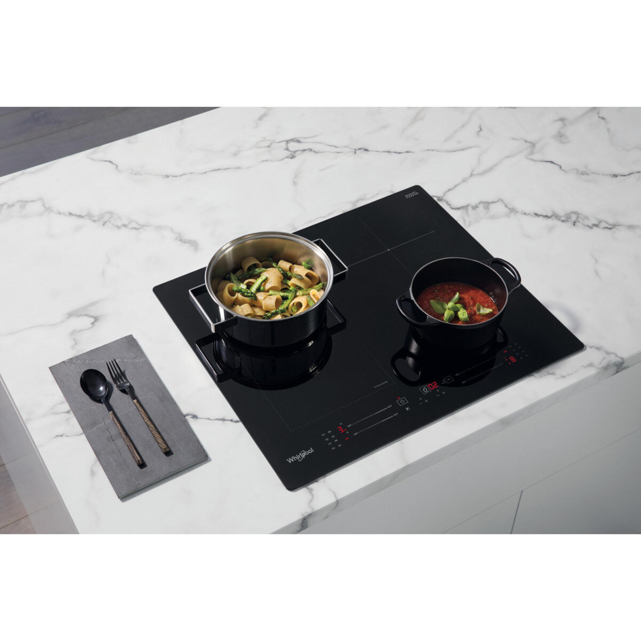 Placa de inducción negra con dos ollas: una con pasta y espárragos, otra con salsa de tomate. Cubiertos sobre tapete gris.