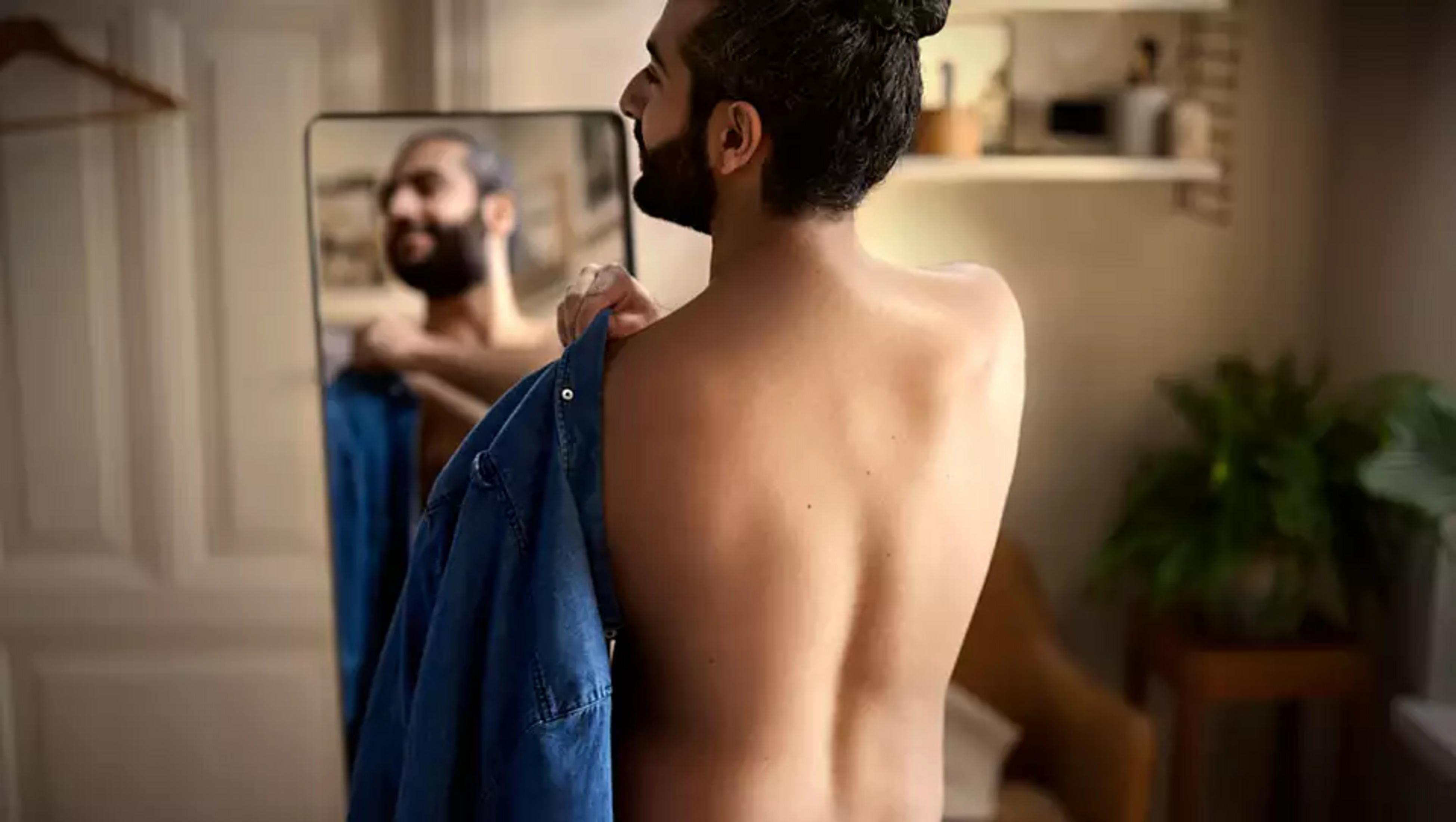 Hombre en espejo poniéndose camisa azul. Vista trasera.