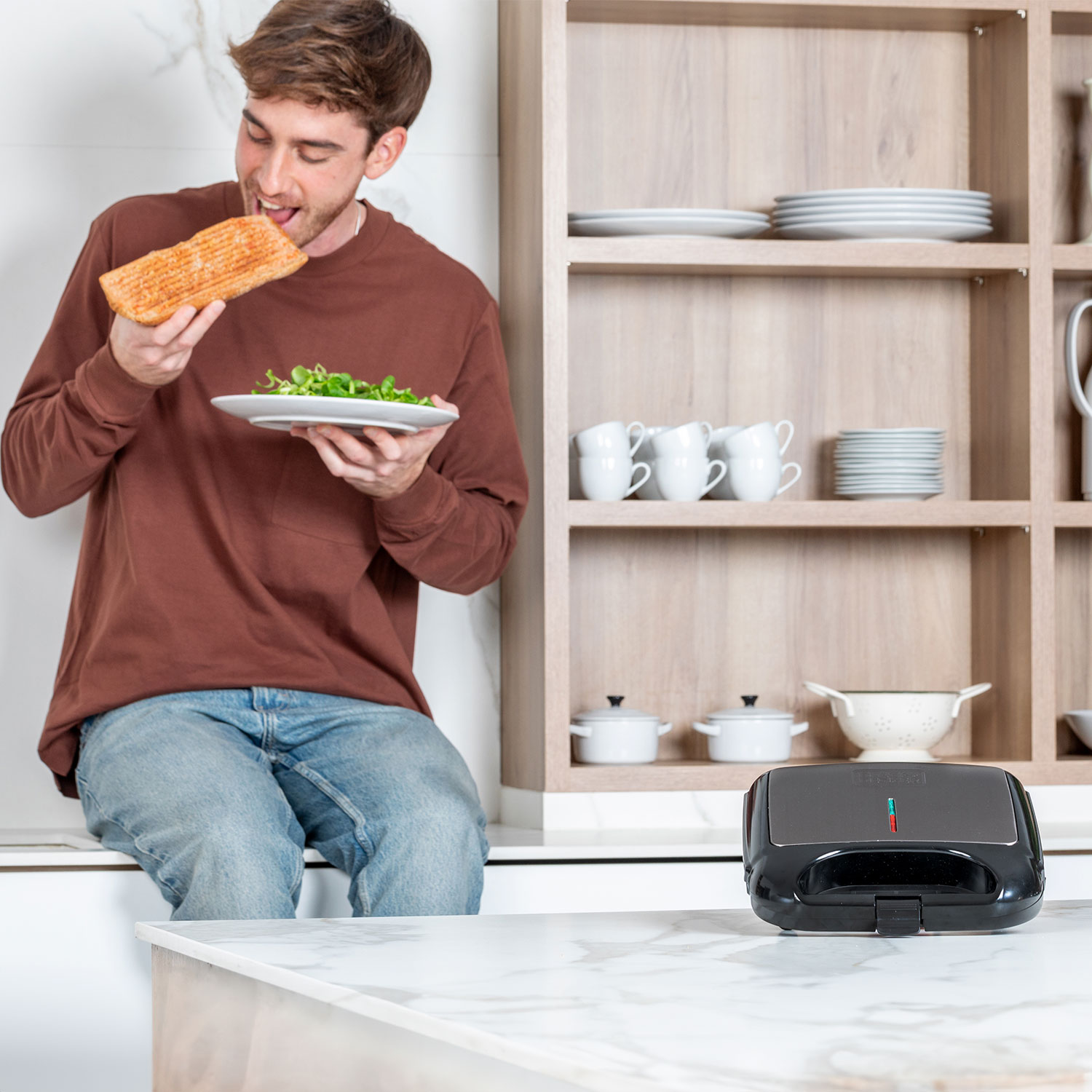 Mężczyzna w brązowej koszuli i dżinsach je grillowaną kanapkę, trzyma sałatkę.