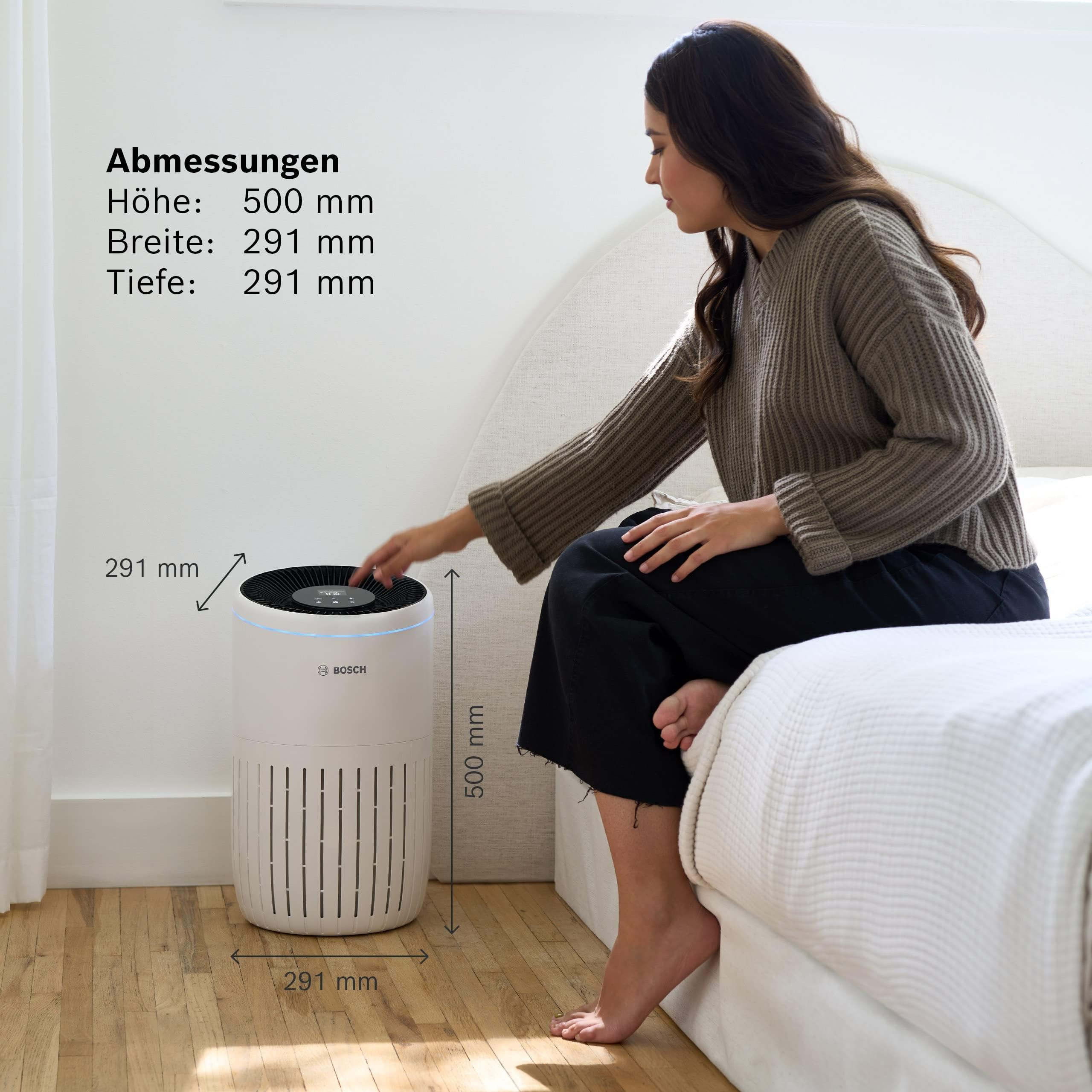 Ein weißer Bosch Luftreiniger steht auf einem Holzboden neben einem Bett. Eine Frau berührt ihn.