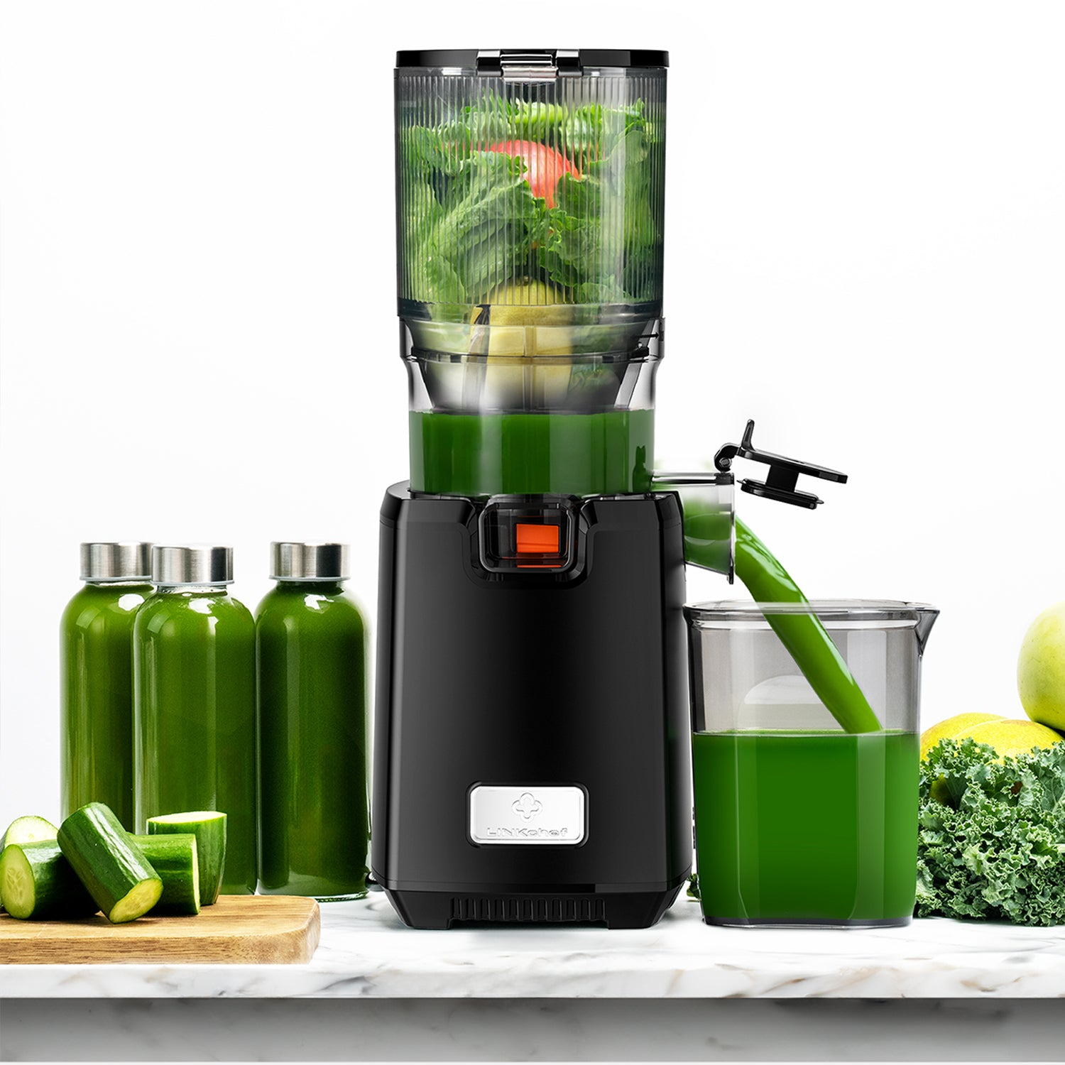 Un estrattore di succo nero con succo verde erogato in una caraffa trasparente. Vicino ci sono diverse bottiglie di succo, cetrioli e cavolo riccio.